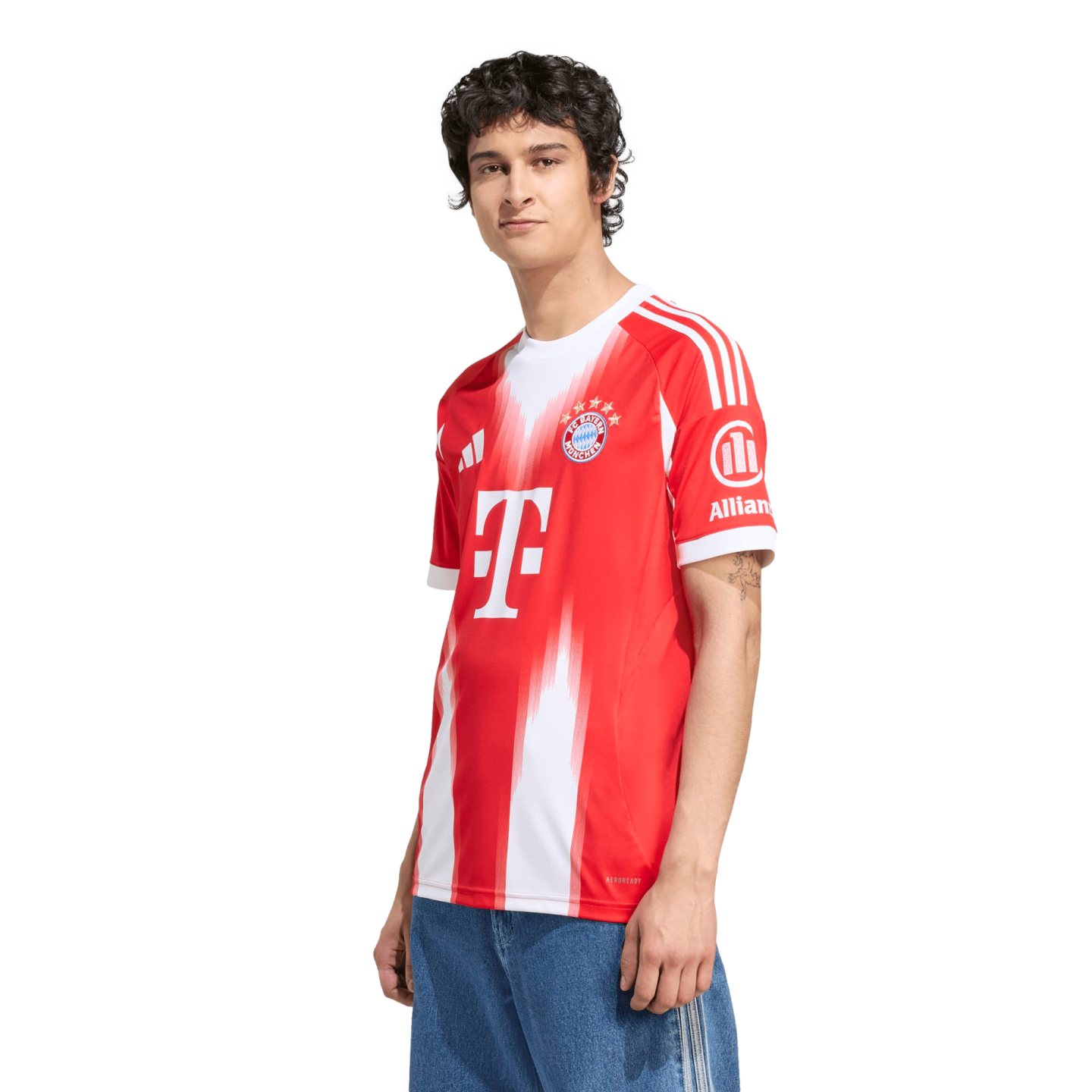A young man with dark curly hair stands against a white background, wearing a red and white Bayern Munich soccer jersey and blue jeans.