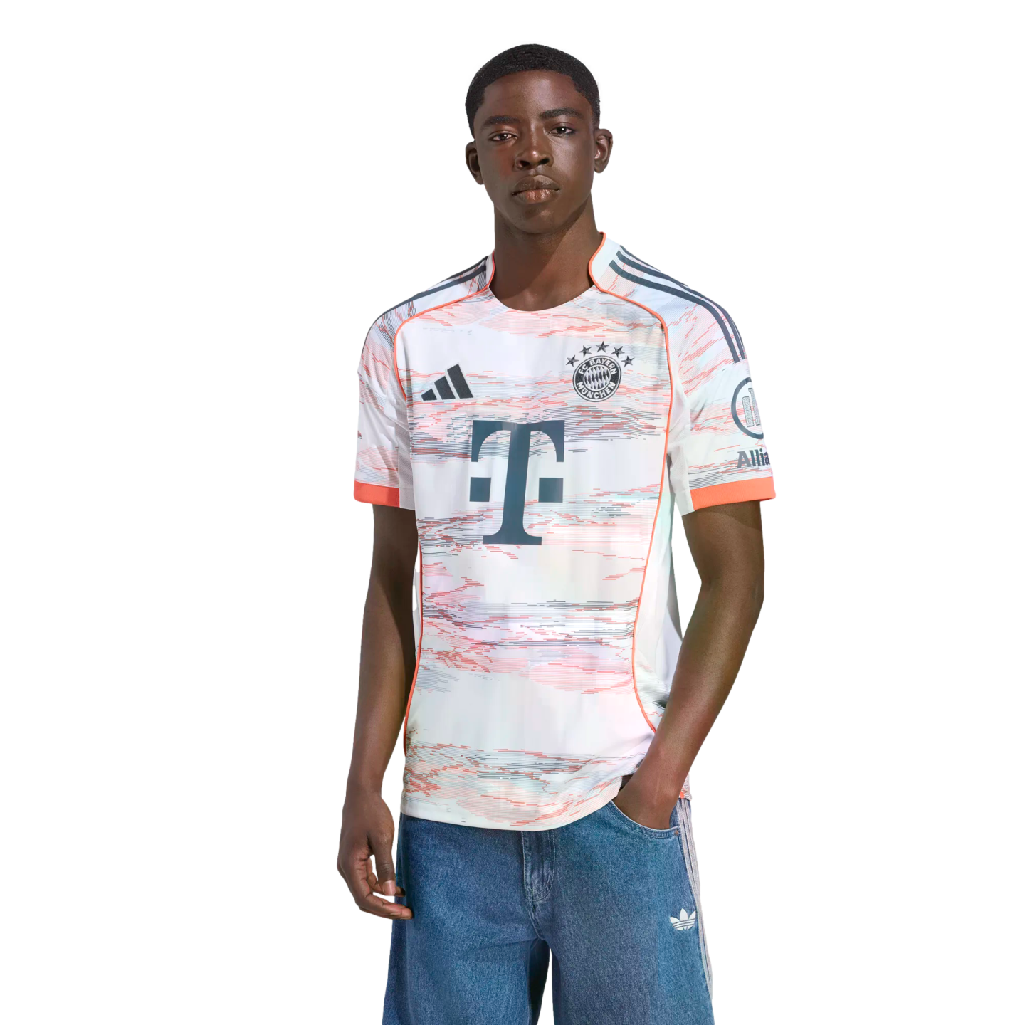 Young Black man modeling a white FC Bayern Munich jersey with a red and grey abstract pattern and blue denim shorts against a white background.
