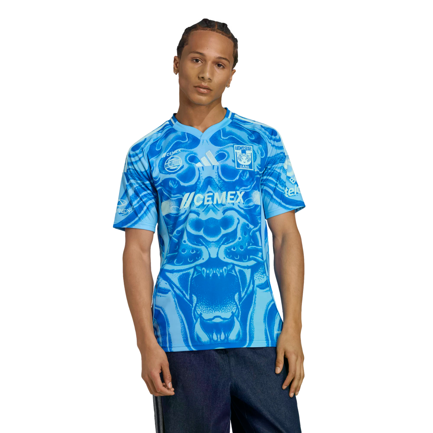 A man in a light blue Tigres UANL soccer jersey with a dark blue stylized tiger graphic on the front and dark jeans, set against a black background.