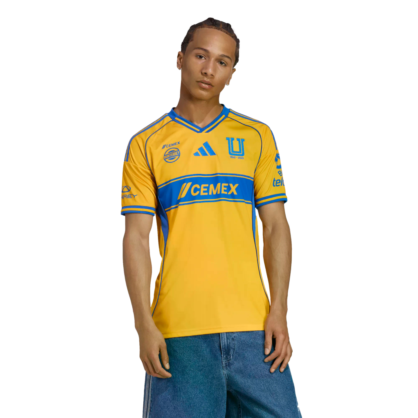 A man models a yellow and blue Tigres UANL soccer jersey with denim shorts against a white background.