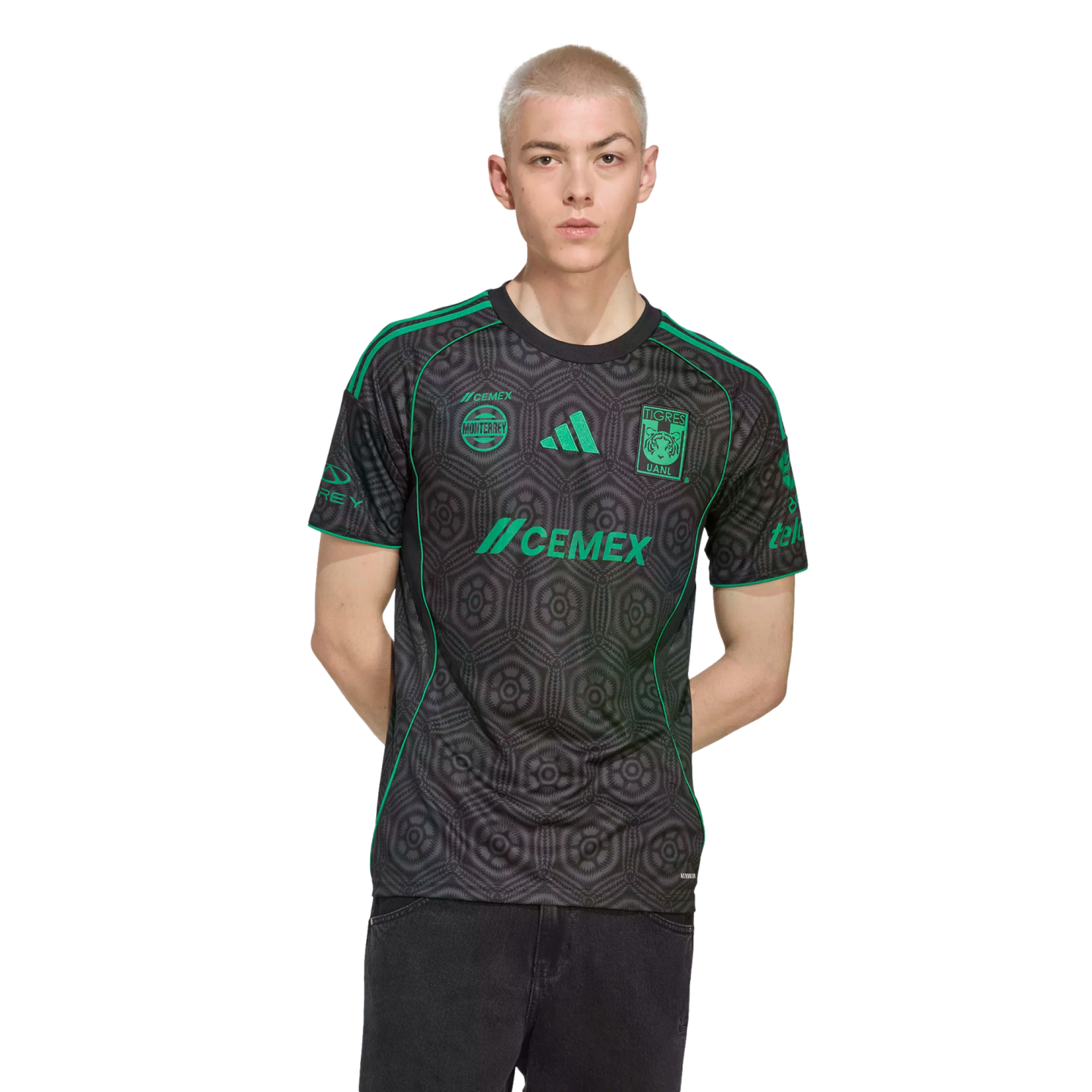 A young man with short blonde hair wearing a black and green Tigres UANL soccer jersey and black jeans, standing against a black background.