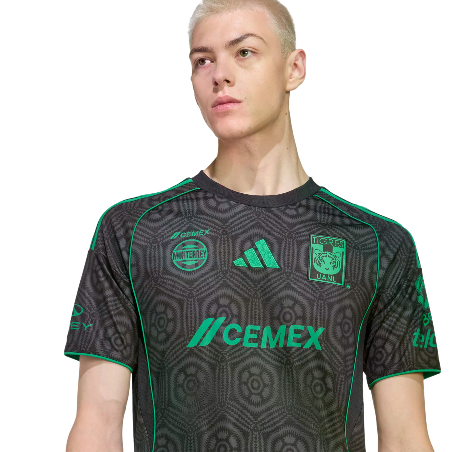 A person with short blonde hair wearing a grey and green patterned Tigres UANL soccer jersey against a black background.