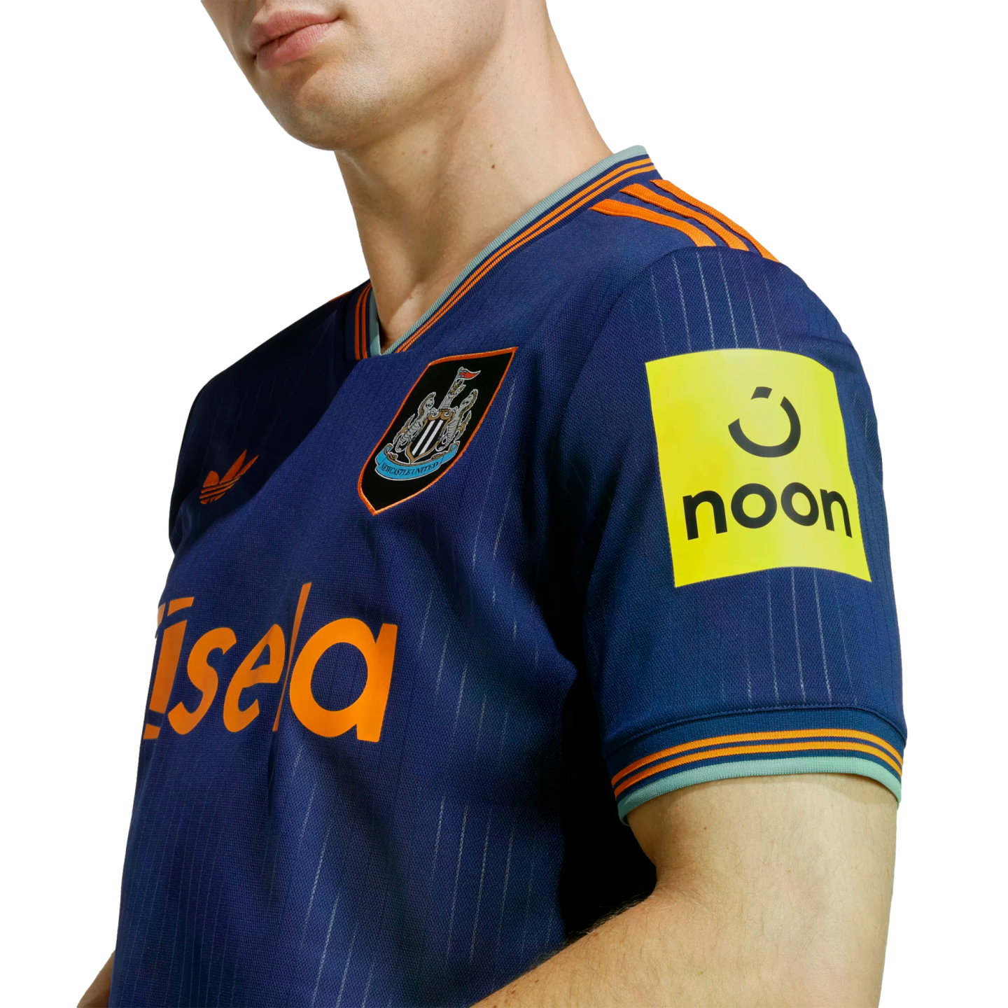A person wears a blue Newcastle United football jersey with orange accents, featuring the club crest, the "Sela" sponsor on the front, and the "noon" logo on the sleeve.