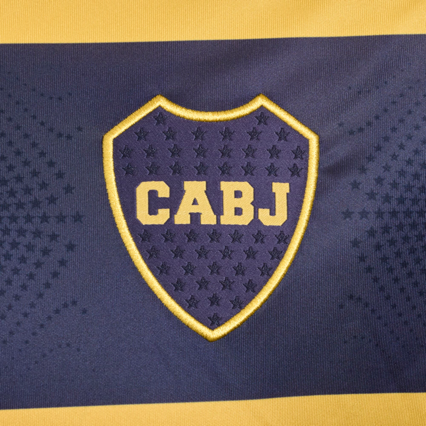 Embroidered Boca Juniors soccer crest with yellow "CABJ" text on a blue shield, set against a blue and yellow striped background.