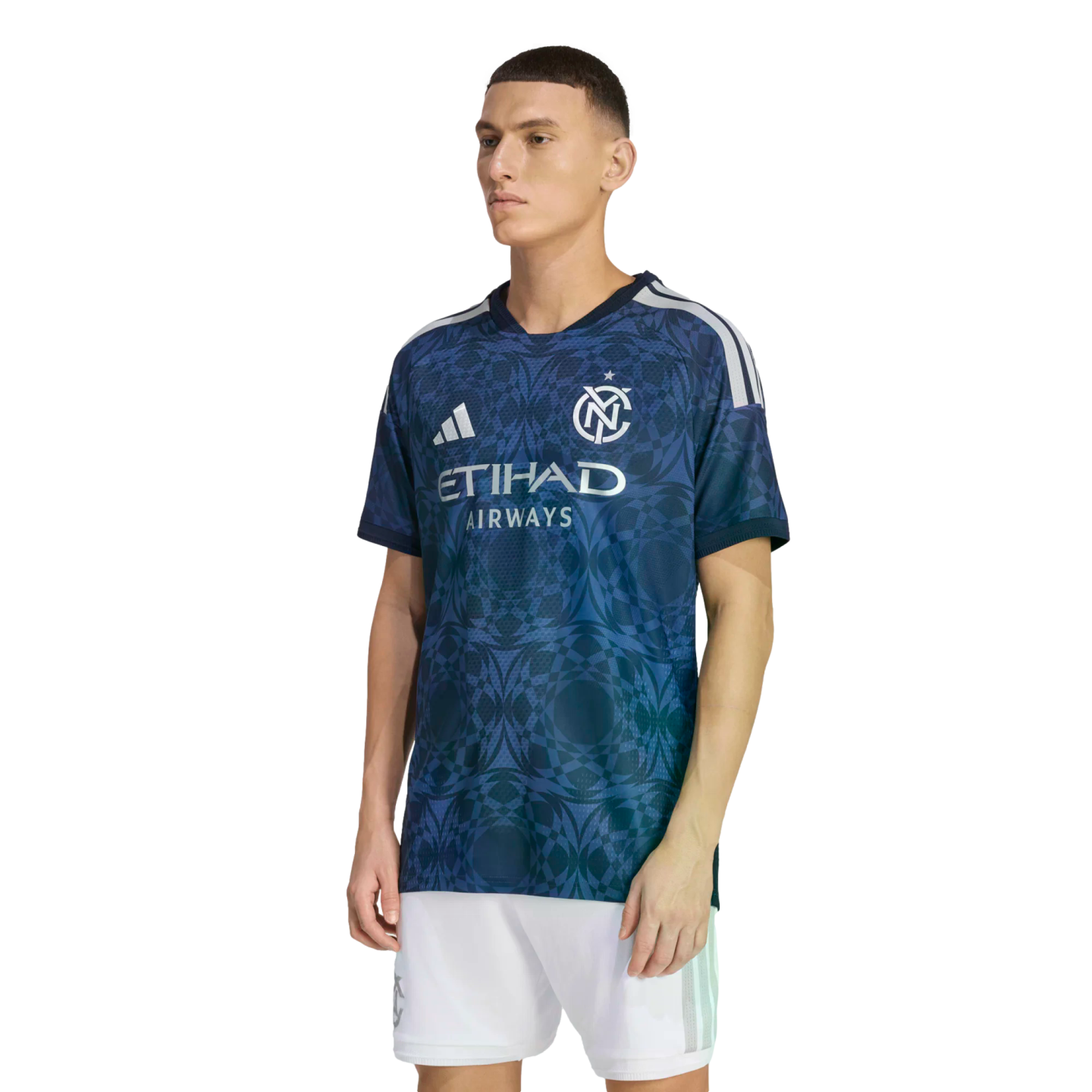 A man models a navy blue NYCFC soccer jersey with a geometric pattern, Adidas logo, and Etihad sponsor, paired with white shorts against a black background.