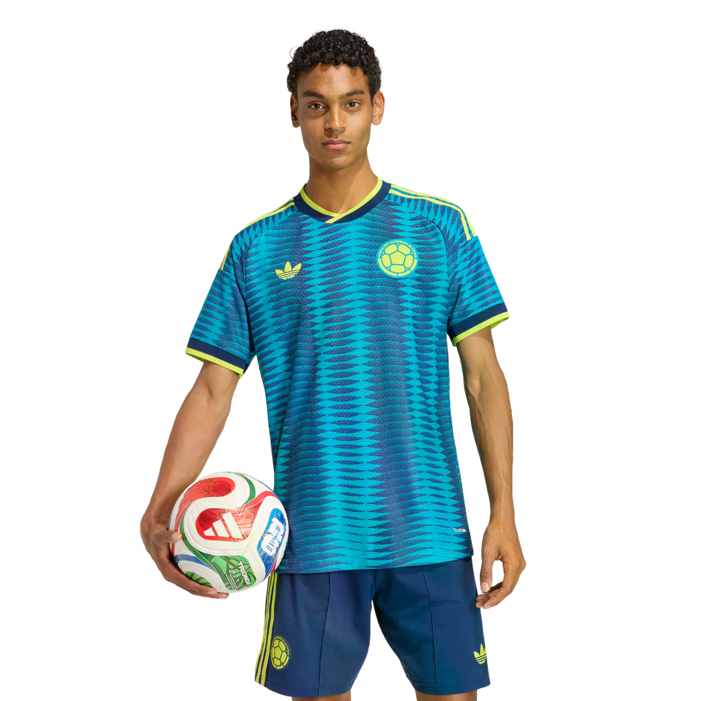 A man in a blue and teal patterned soccer jersey and matching shorts holds a colorful soccer ball against a solid black background.