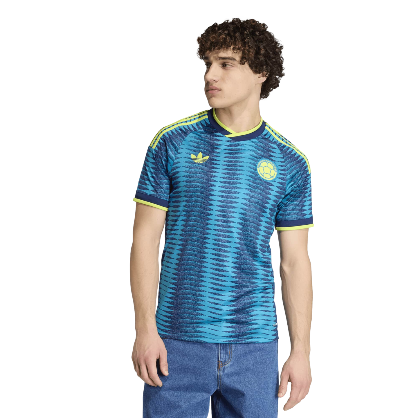 Young man with curly hair wearing a blue patterned soccer jersey and blue jeans, standing against a black background.