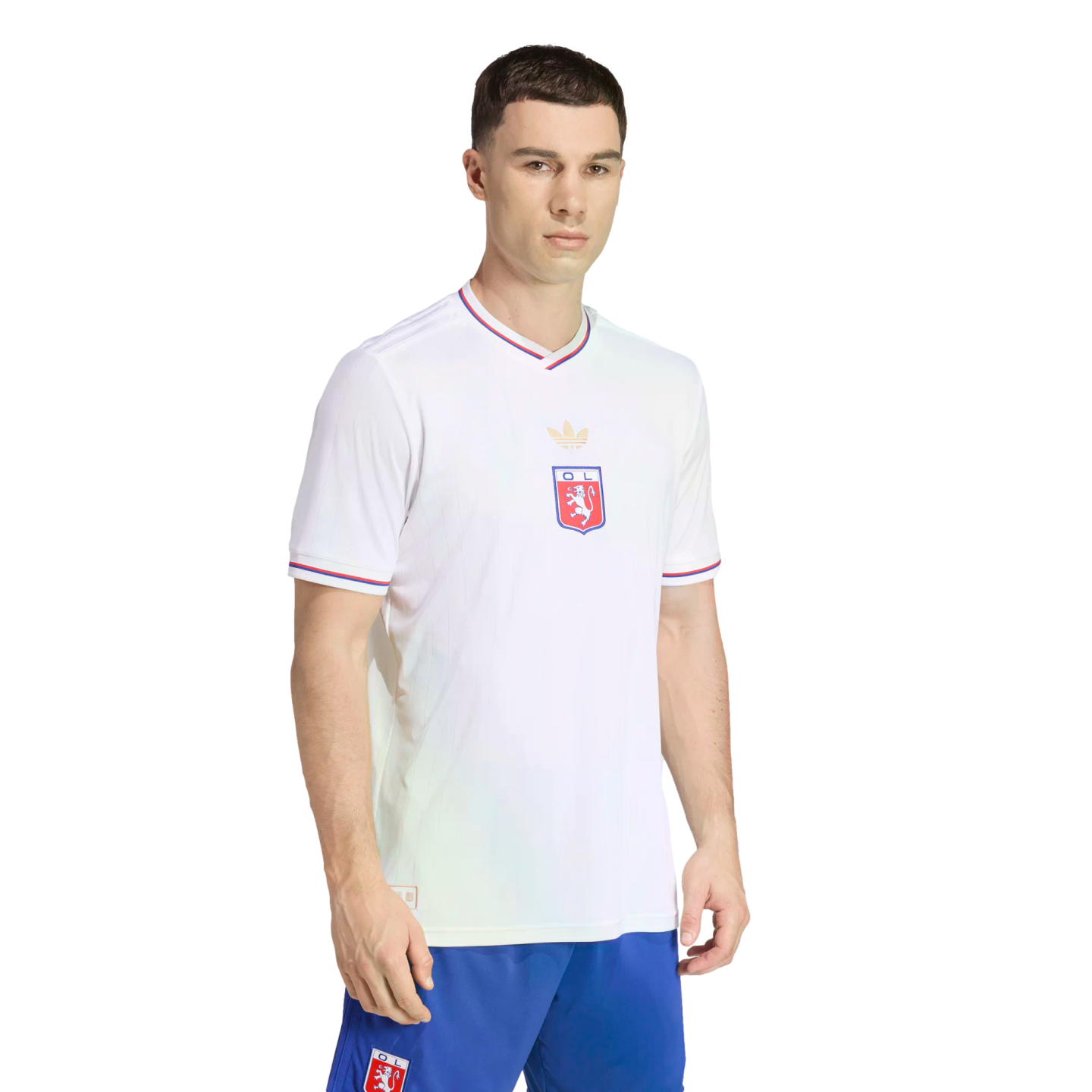 A man models a white Olympique Lyonnais soccer jersey with a gold trefoil logo and blue and red trim, paired with blue shorts, against a solid black background.