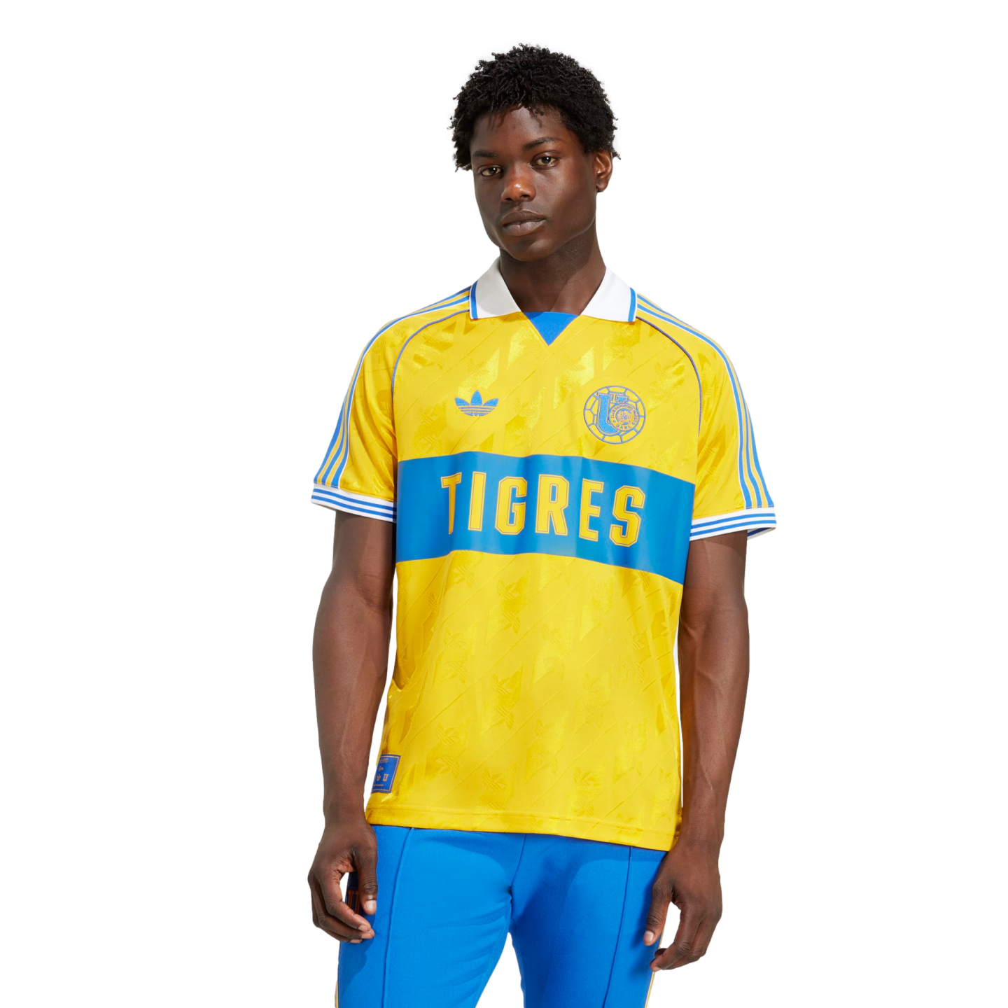 A young man with dark skin wears a yellow and blue Tigres UANL soccer jersey and blue pants against a solid black background.