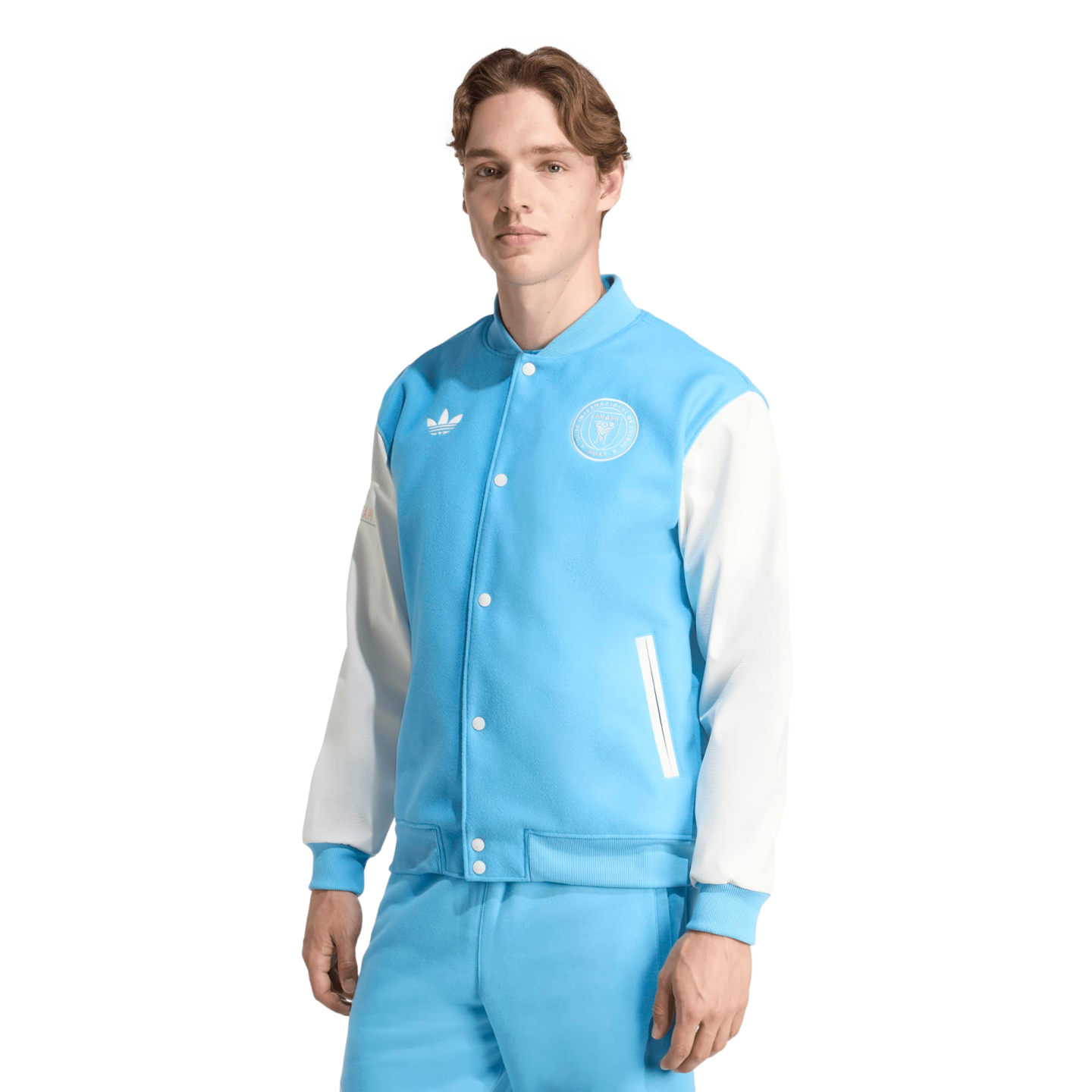 A man models a light blue and white Adidas varsity jacket with an Inter Miami crest and matching light blue pants against a black background.