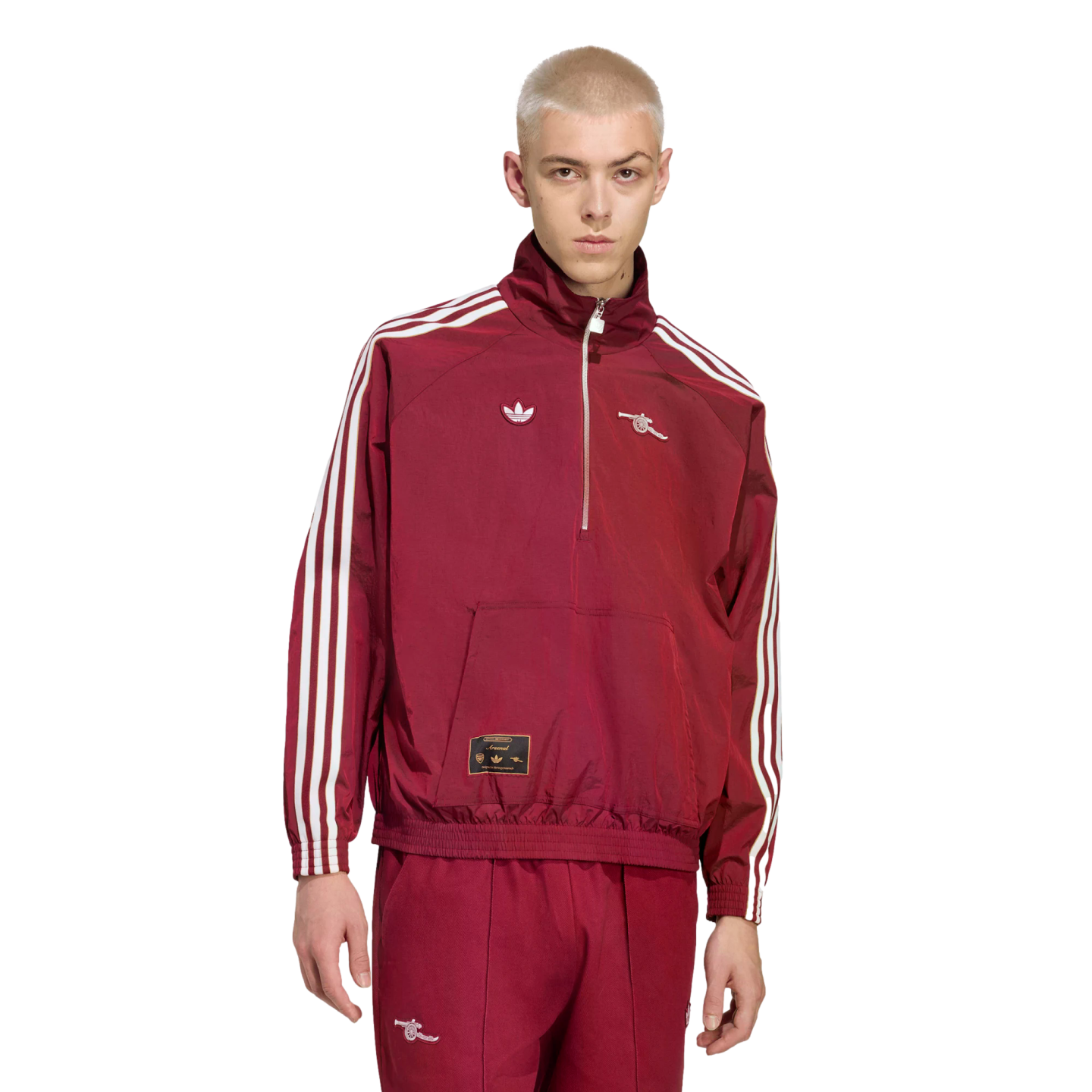 A person with short blonde hair models a maroon Adidas x Arsenal tracksuit with white stripes and logos against a solid black background.