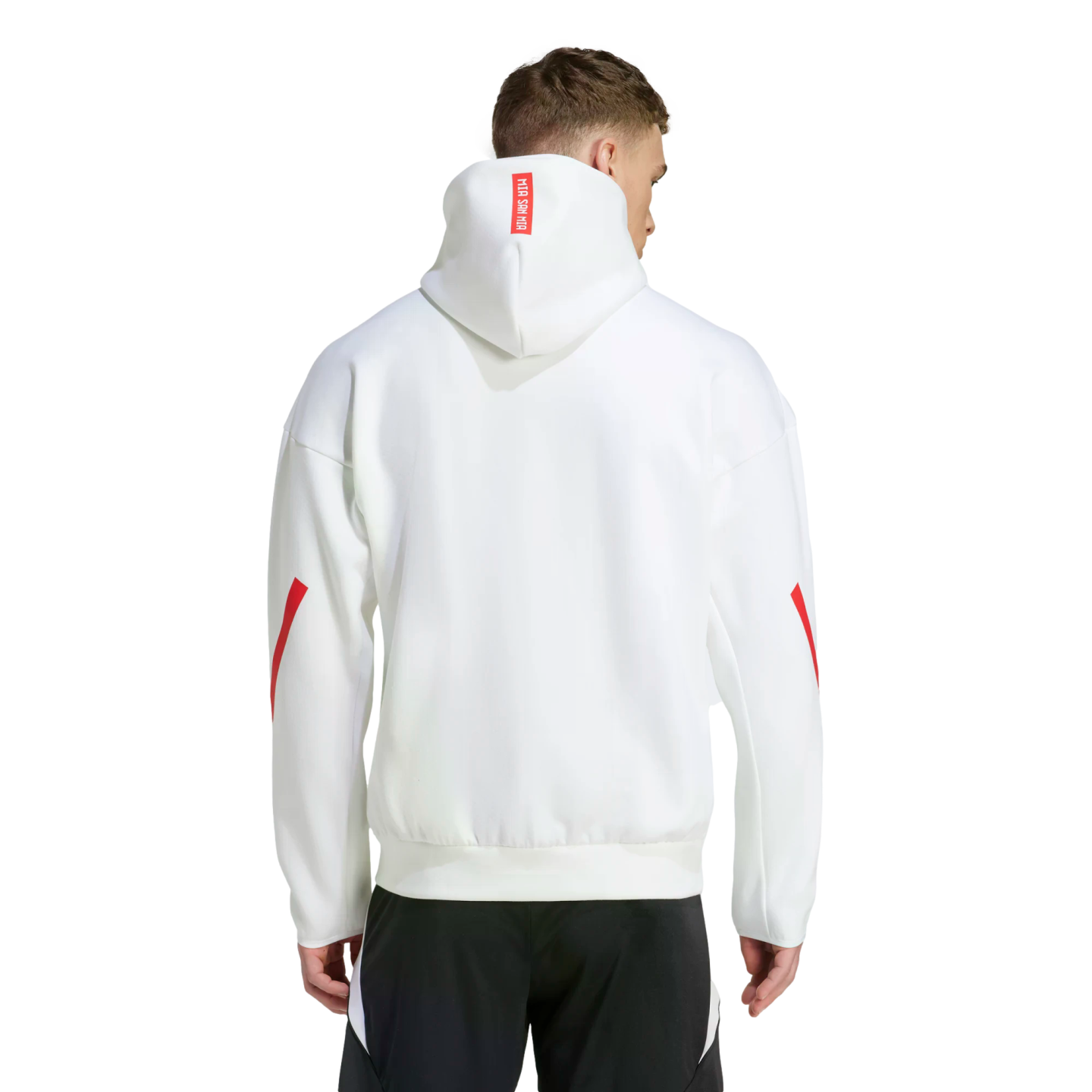 Rear view of a man wearing a white hoodie with red sleeve stripes and a red "MIA SAN MIA" label on the hood, paired with black athletic pants.