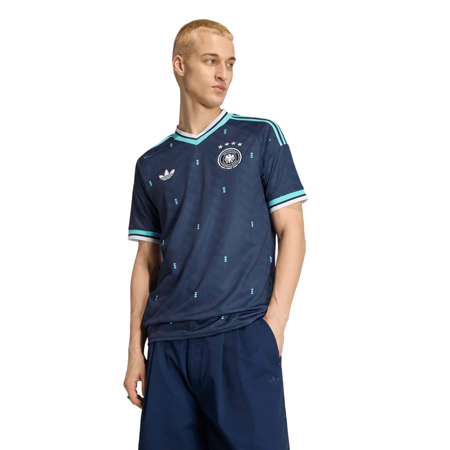 A young man with blonde hair wearing a dark blue Adidas Germany national team soccer jersey and navy pants, standing against a solid black background.