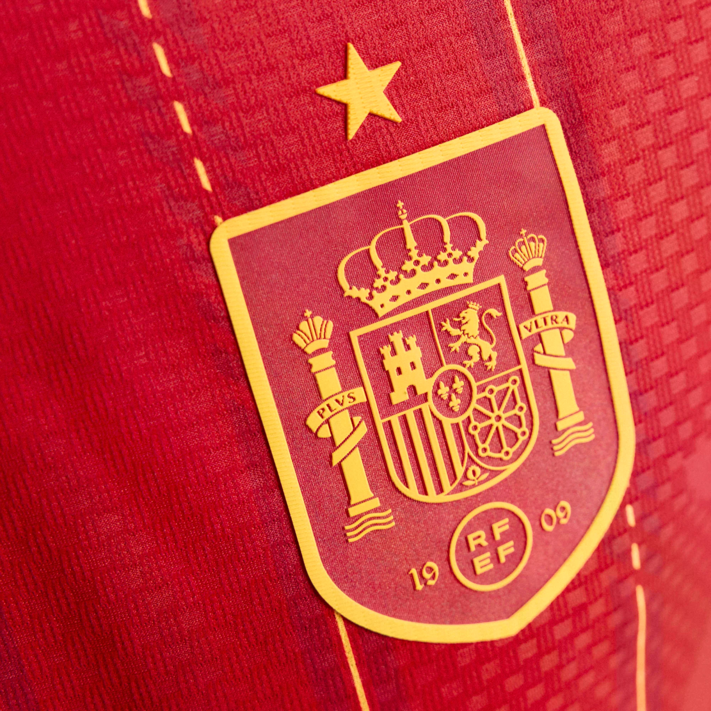 A close-up of the yellow Spanish national football team crest and star on a red textured jersey.