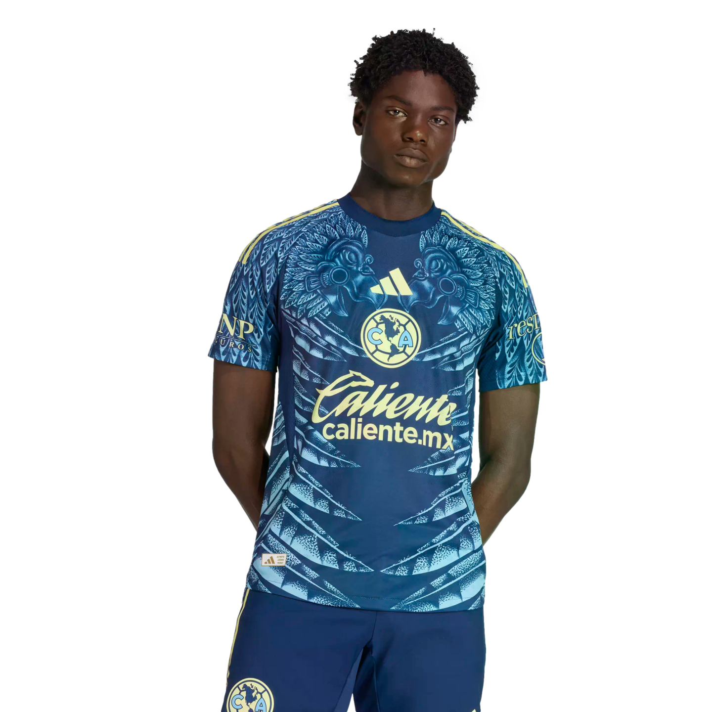A Black man wearing a blue and yellow Club América soccer kit featuring intricate feather patterns and sponsor logos, posed against a plain black background.