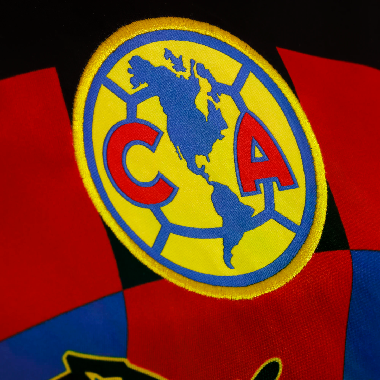 Close-up of an embroidered Club América logo on patterned fabric. The yellow patch features a blue map of the Americas flanked by a red 'C' and 'A'.