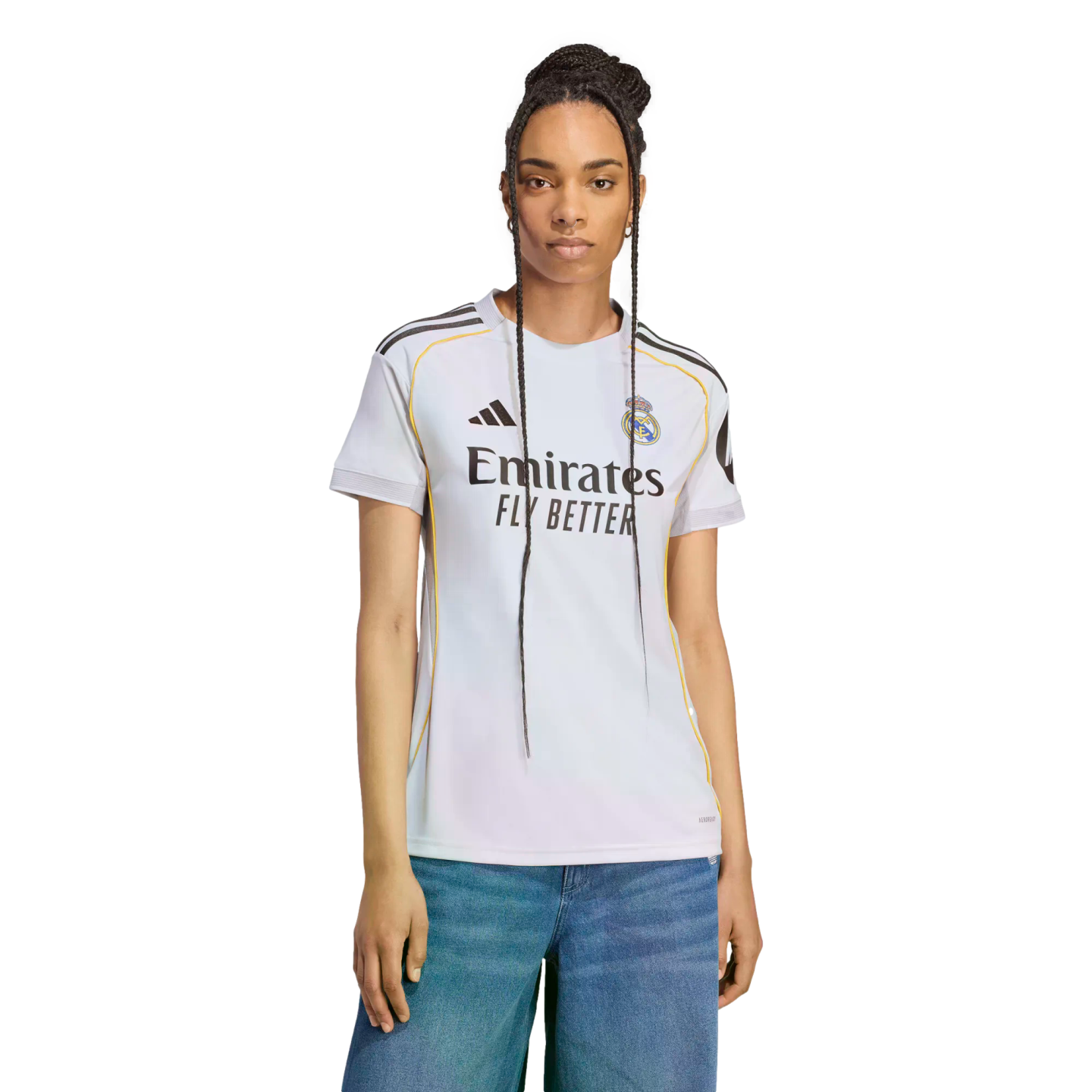 A woman with long braids poses in a white Real Madrid soccer jersey and blue jeans against a white background.