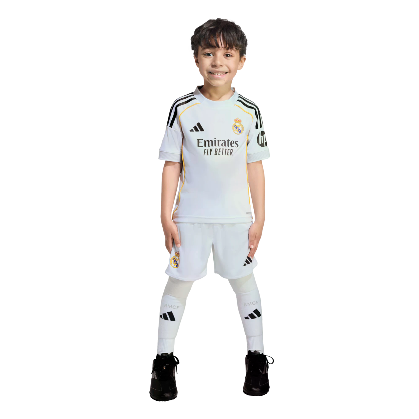 Smiling young boy in a full white Real Madrid football kit and black shoes, standing against a plain white background.