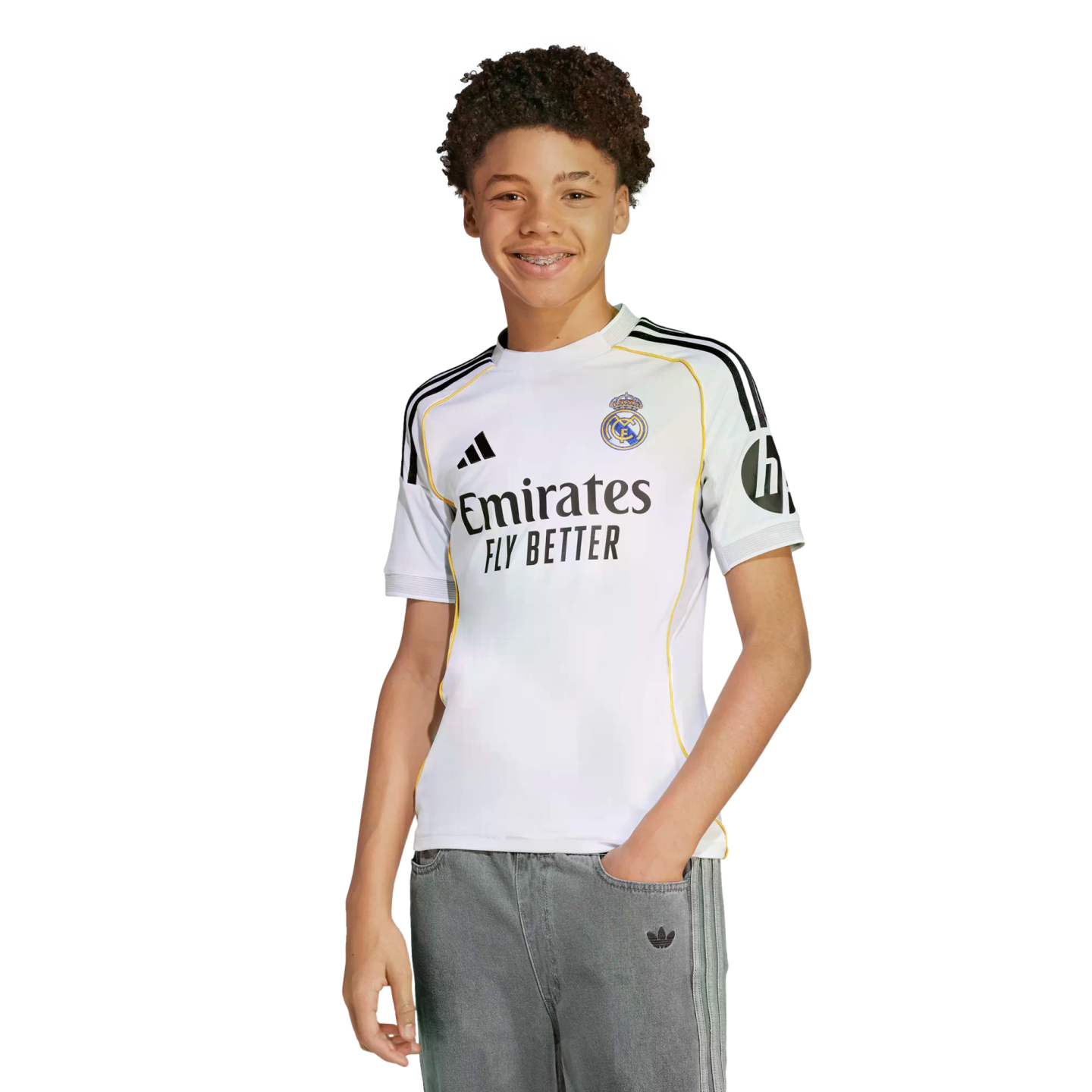 Smiling young person with curly hair and braces, wearing a white Real Madrid jersey and grey pants, isolated on a white background.