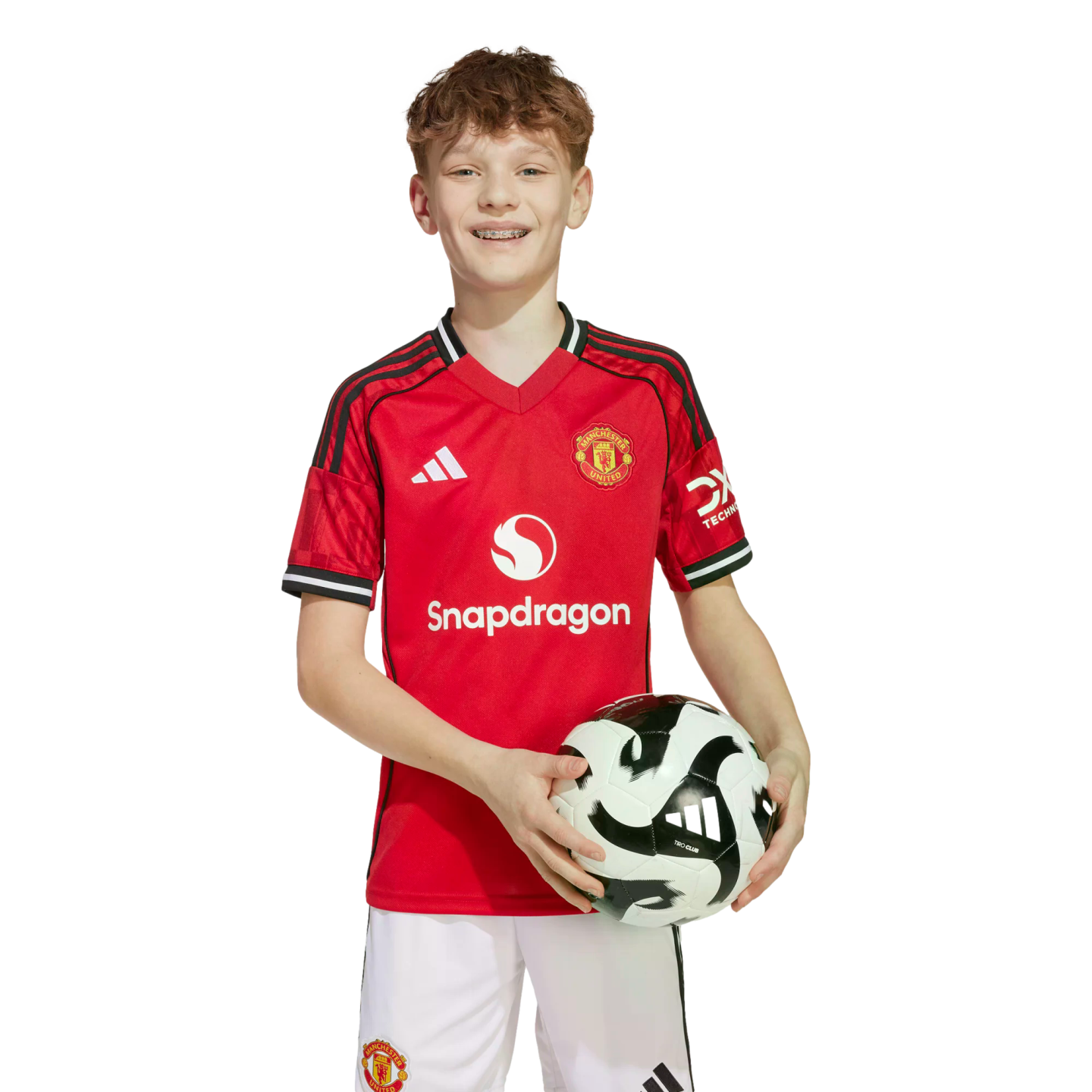 A smiling young boy with braces wearing a red Manchester United soccer jersey and white shorts while holding a soccer ball against a white background.