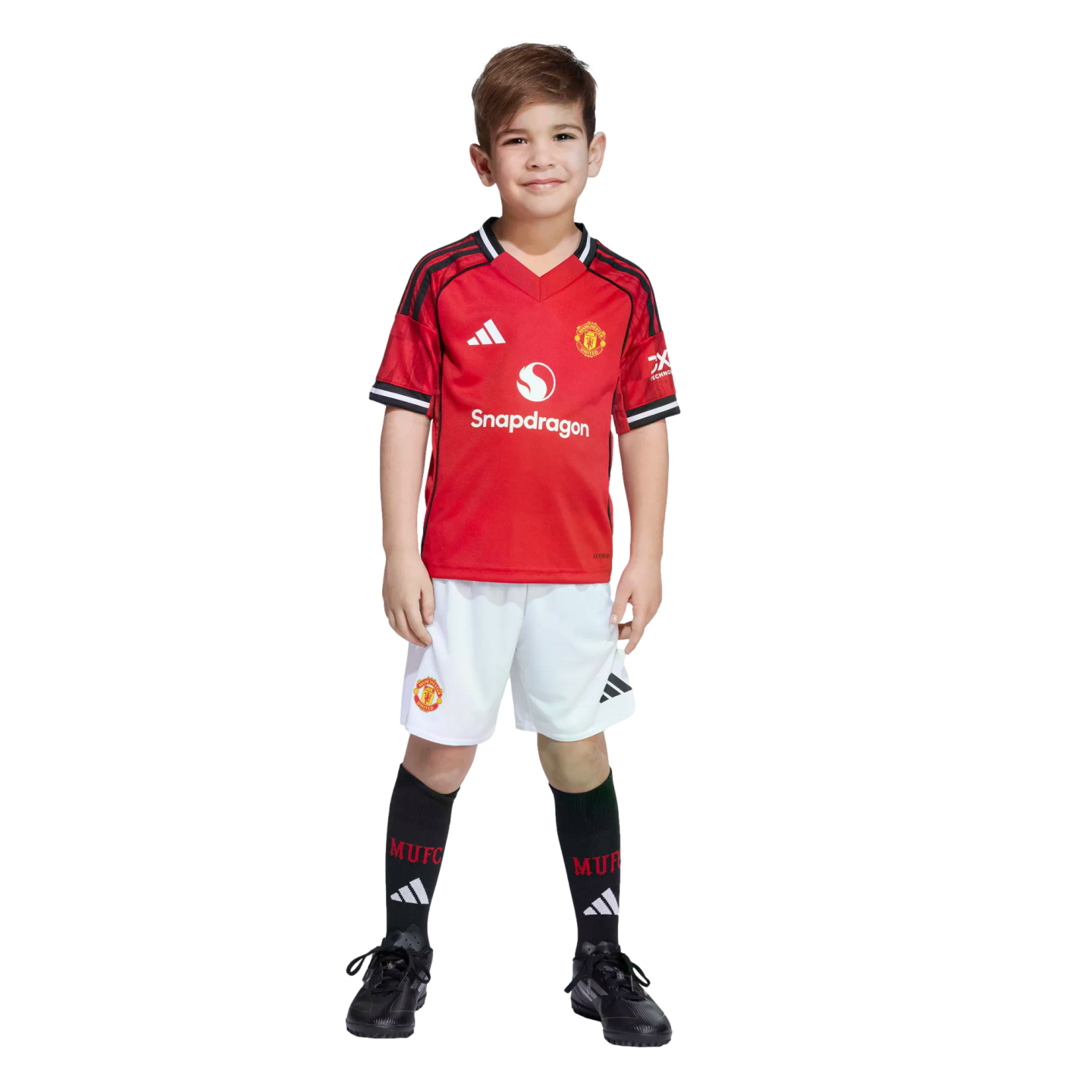 A young boy smiles while wearing a full Manchester United football kit, featuring a red jersey with a Snapdragon logo, white shorts, and black socks.