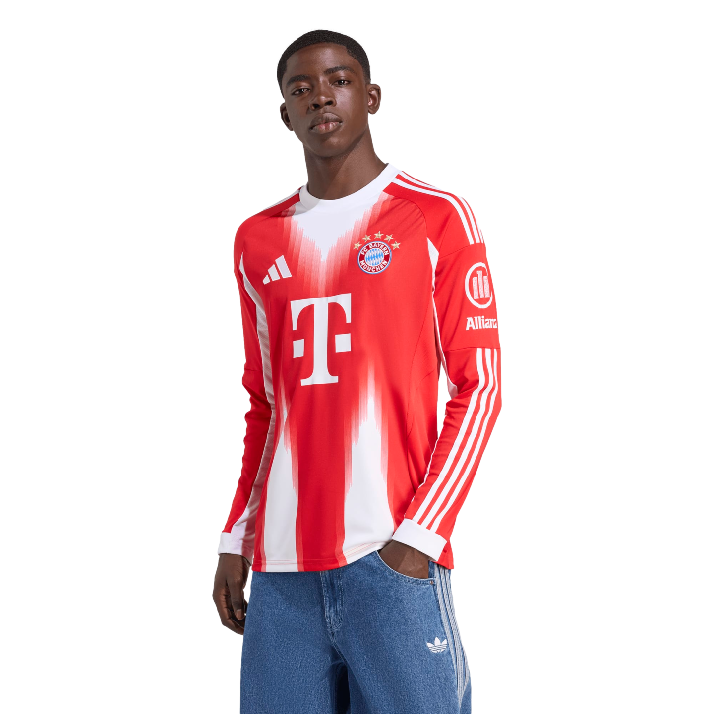 A Black man wearing a long-sleeved red and white Adidas FC Bayern Munich soccer jersey and blue jeans, standing with one hand in his pocket against a white background.