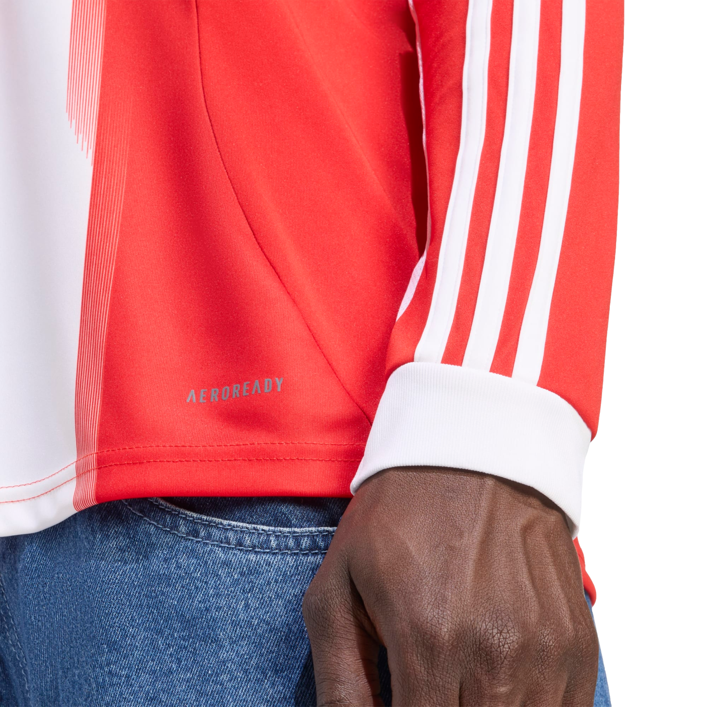 Close-up of a person wearing a red and white long-sleeved shirt with three white stripes on the sleeve, "AEROREADY" branding on the side, and blue denim jeans.