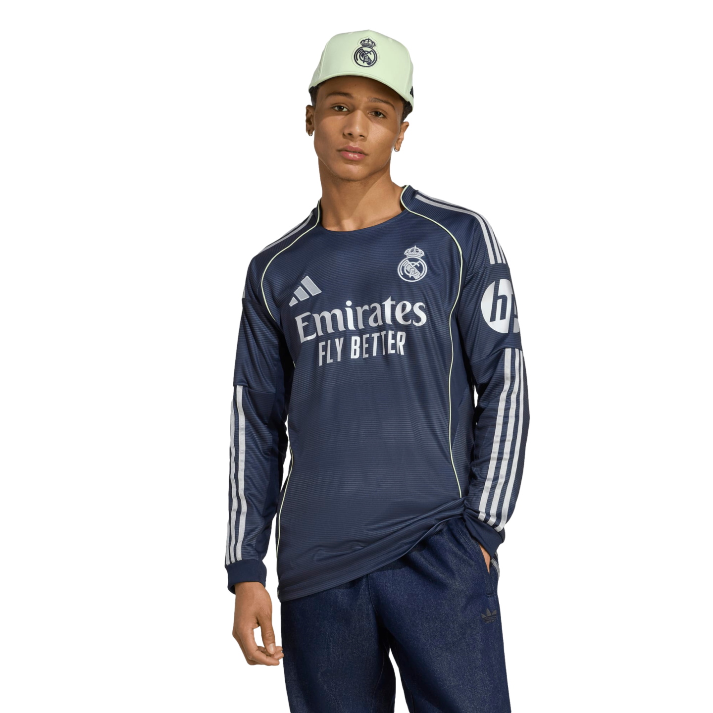 A man posing in a navy blue Real Madrid long-sleeve training shirt, a mint green logo cap, and dark blue trousers against a white background.