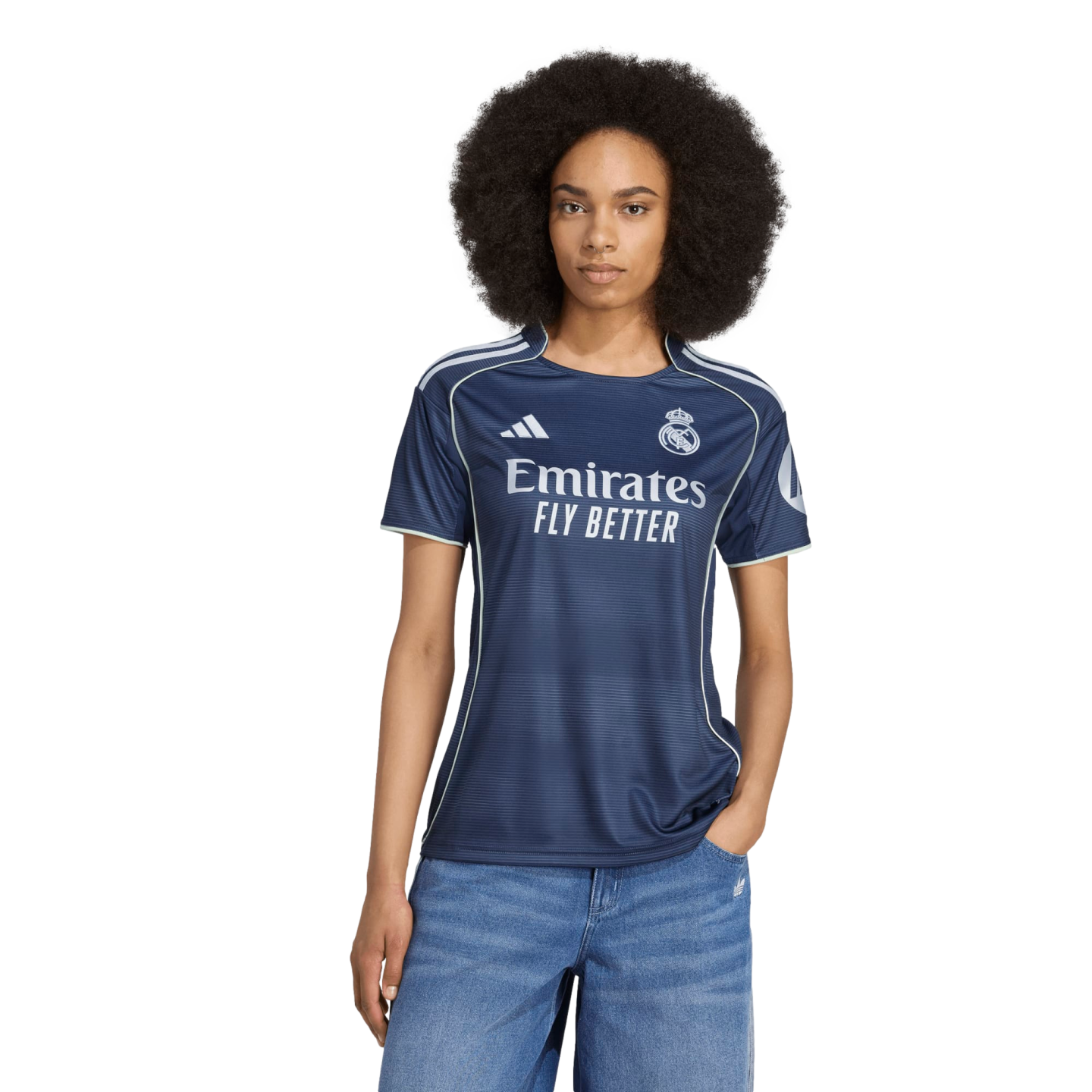 A woman with a large afro wears a navy blue Real Madrid soccer jersey and blue jeans against a white background.