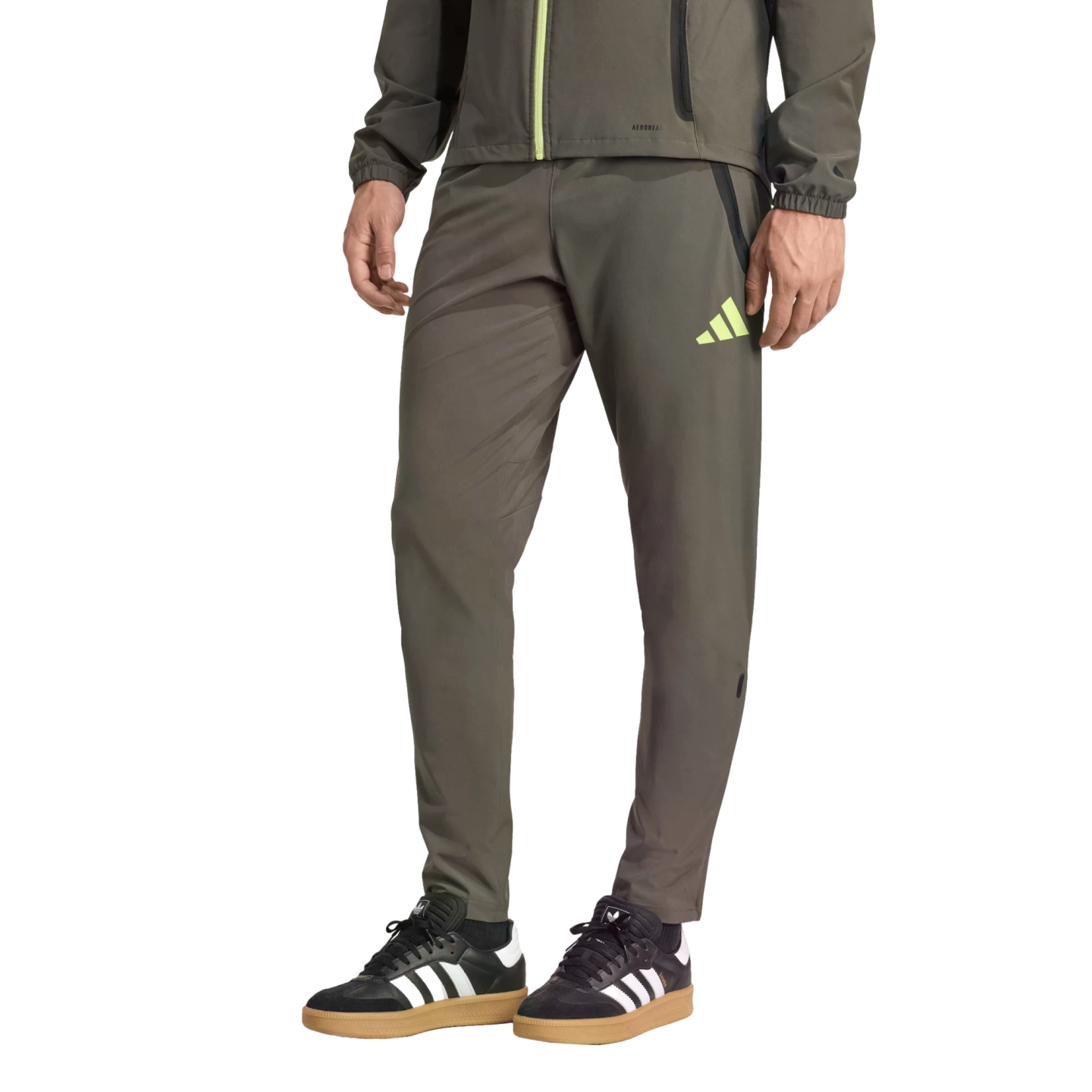 Lower body of a person wearing olive green Adidas track pants and black sneakers with white stripes and gum soles against a black background.
