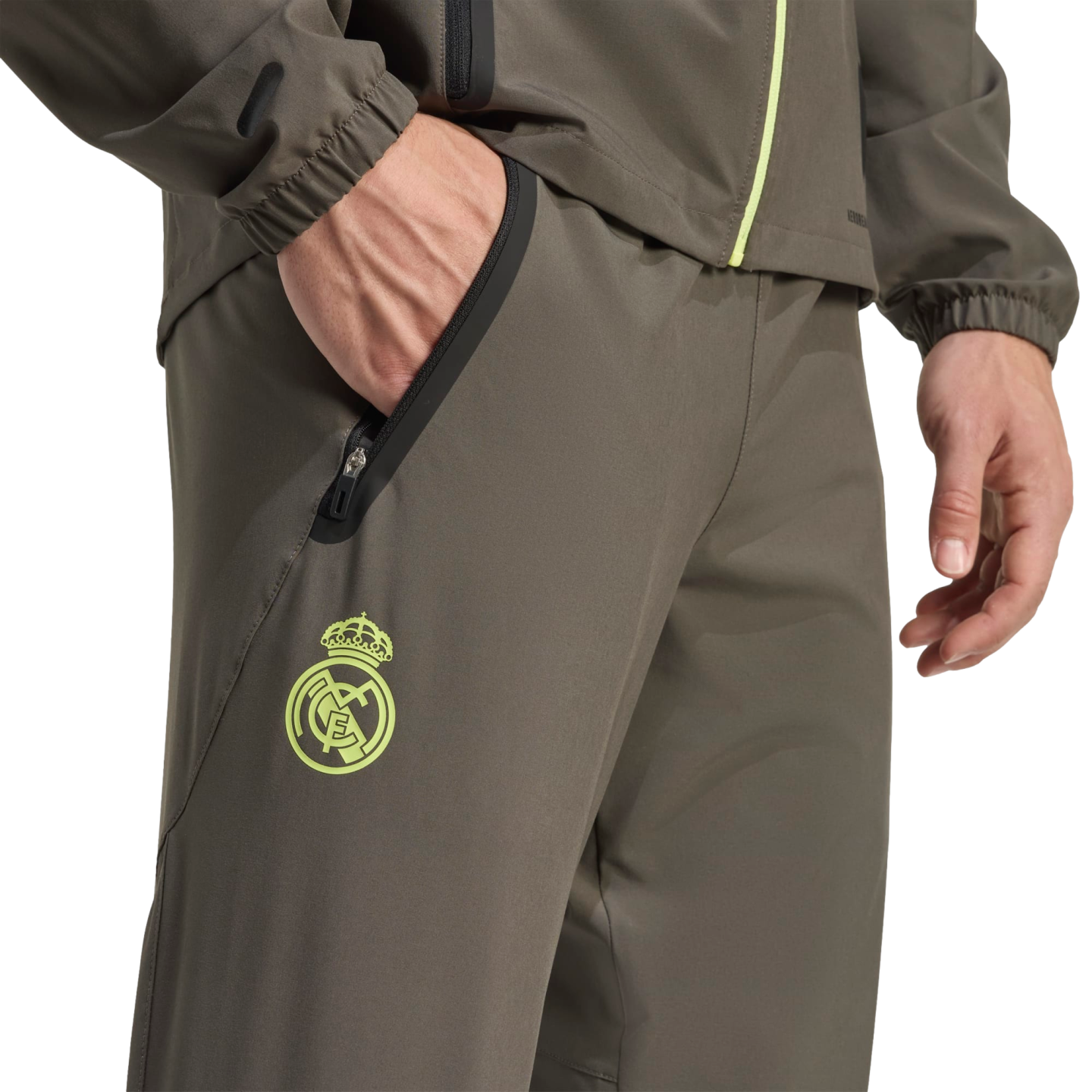 A close-up of a person wearing an olive green tracksuit with a lime green Real Madrid crest and zippered pockets.