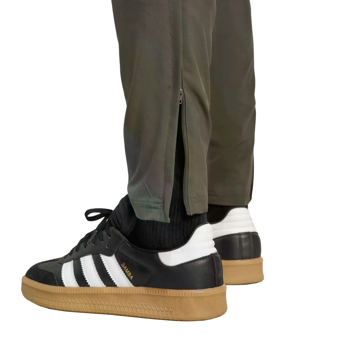 A low-angle view of black Adidas Samba sneakers with gum soles, worn with olive green pants featuring ankle zippers, set against a black background.