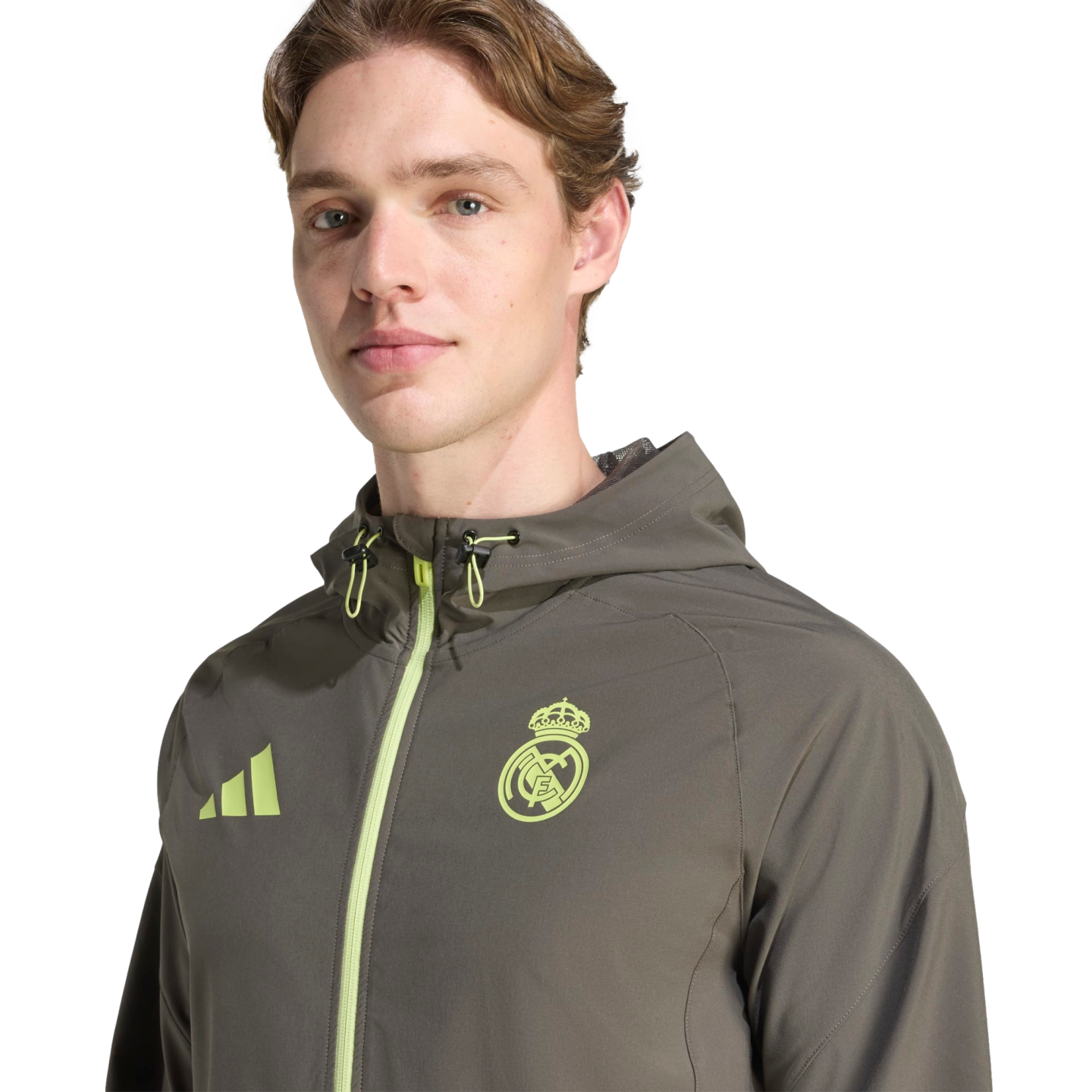 A man wearing an olive green Real Madrid Adidas windbreaker with neon yellow accents, posed against a black background.