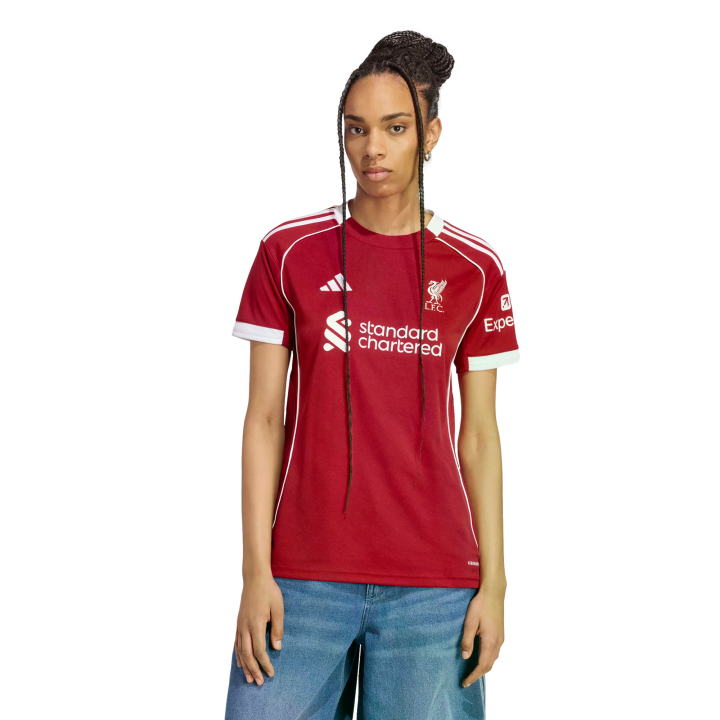 A woman with long braids models a red Liverpool F.C. soccer jersey with white trim and sponsor logos, paired with blue jeans against a black background.
