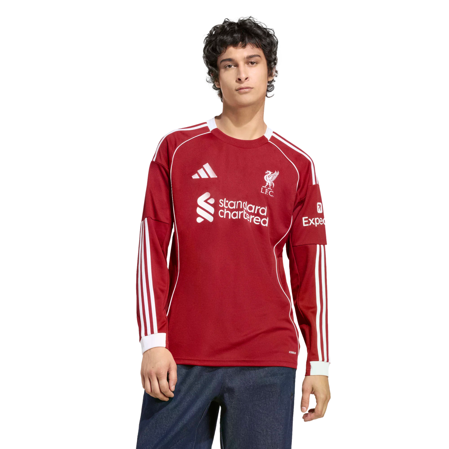 A man models a red long-sleeved Liverpool FC jersey with white accents and sponsor logos, paired with blue jeans, against a black background.