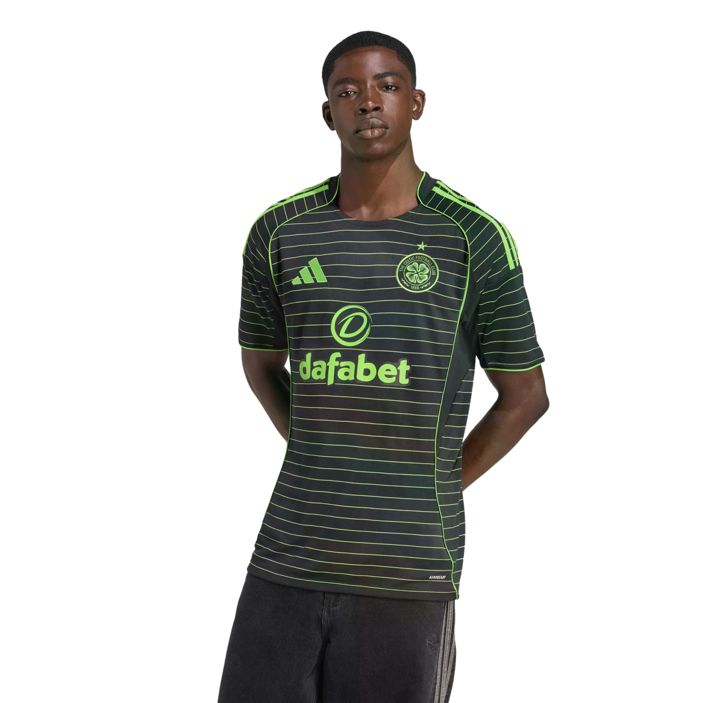 A young man models a black Celtic FC jersey featuring thin neon green horizontal stripes and logos, posed against a solid black background.