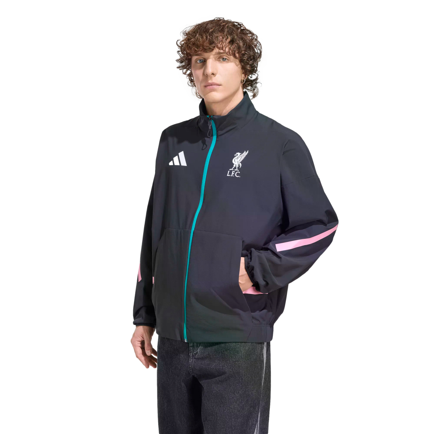 A person with curly brown hair models a black Adidas Liverpool F.C. track jacket featuring teal and pink accents, standing against a solid black background.