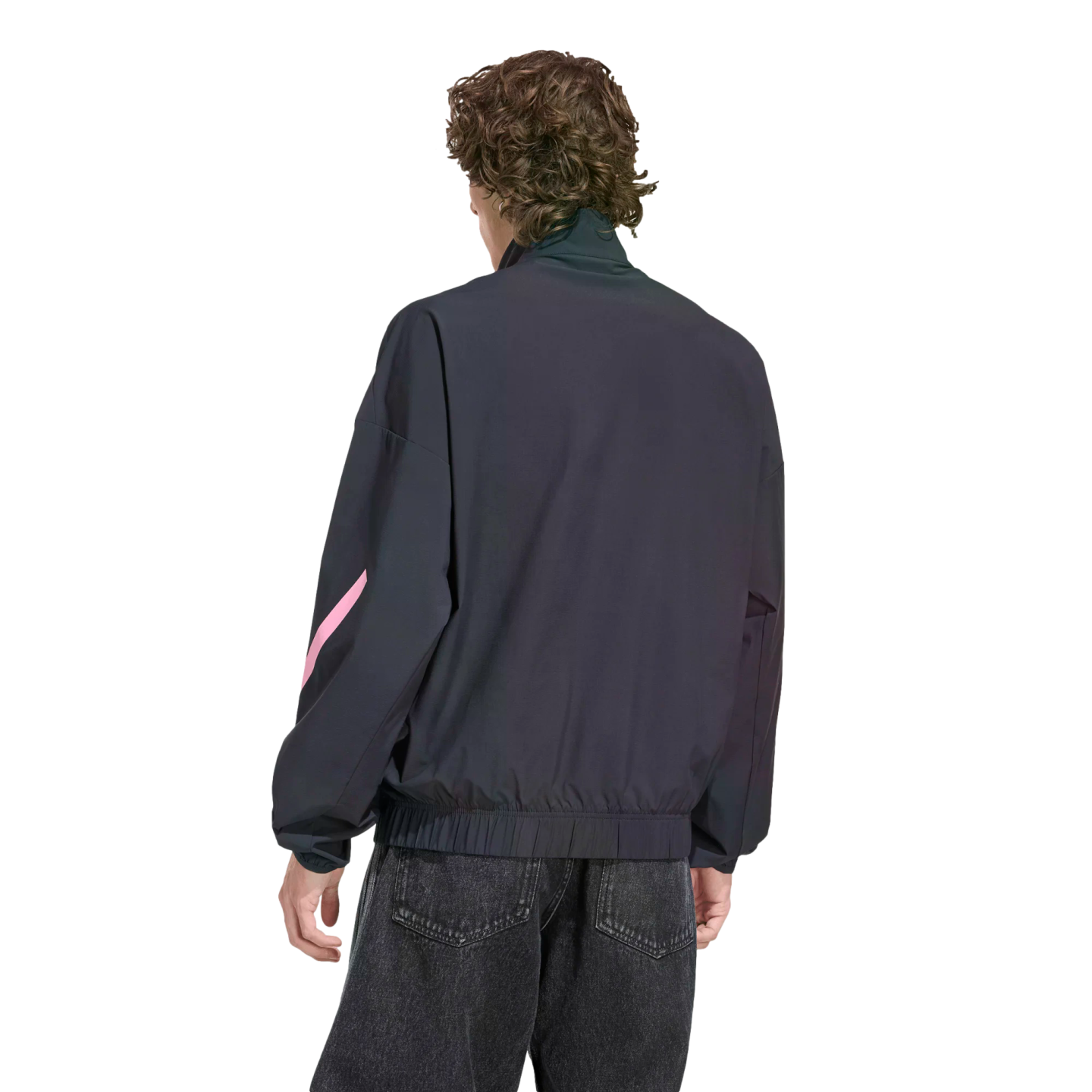 Rear view of a person with wavy brown hair wearing a dark navy zip-up jacket with a pink design on the sleeve and black jeans, set against a black background.