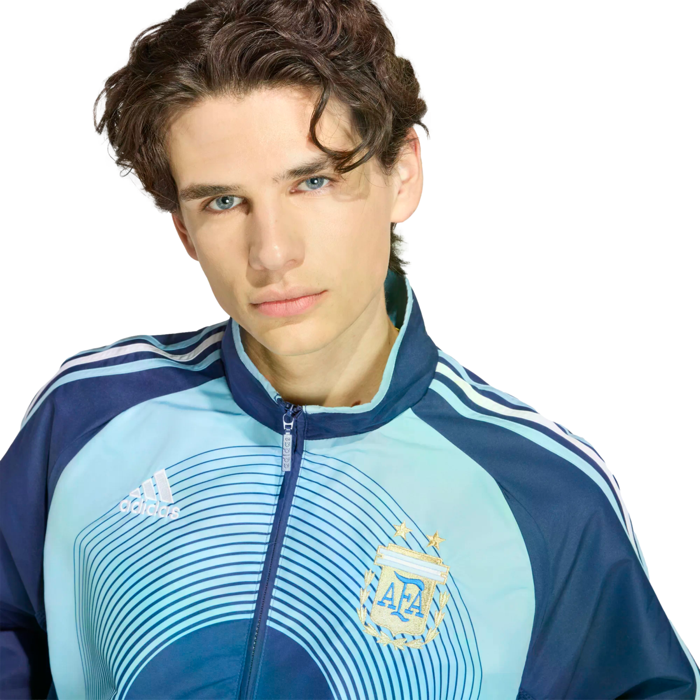 A young man with wavy dark hair and blue eyes wears a blue Argentina national football team track jacket, set against a solid black background.