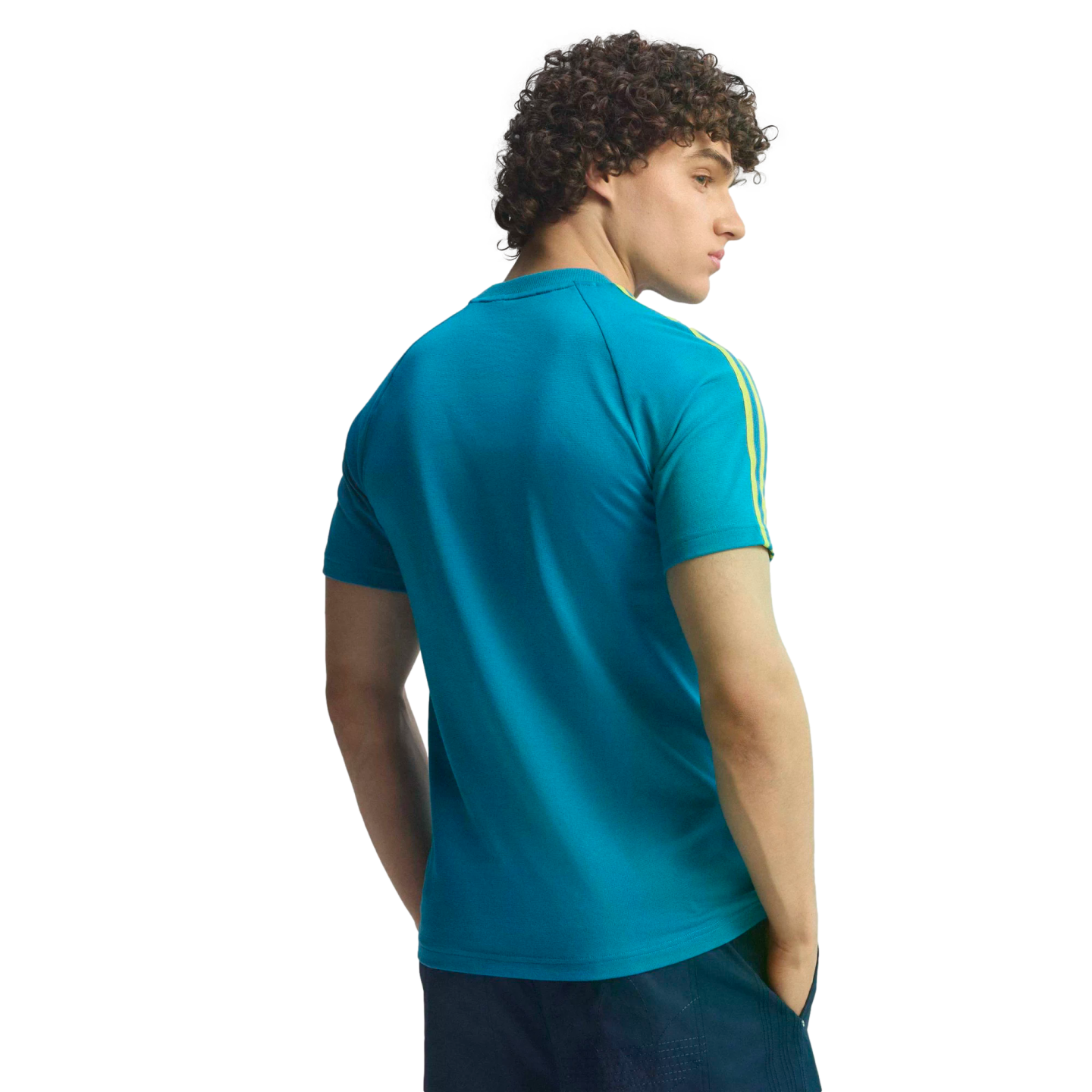 Back view of a man with curly hair in a teal t-shirt with yellow sleeve stripes, looking over his shoulder against a black background.
