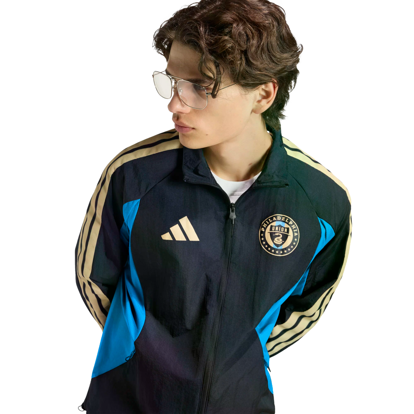 Young person with wavy hair and glasses wearing a black, blue, and gold Philadelphia Union Adidas track jacket against a black background.