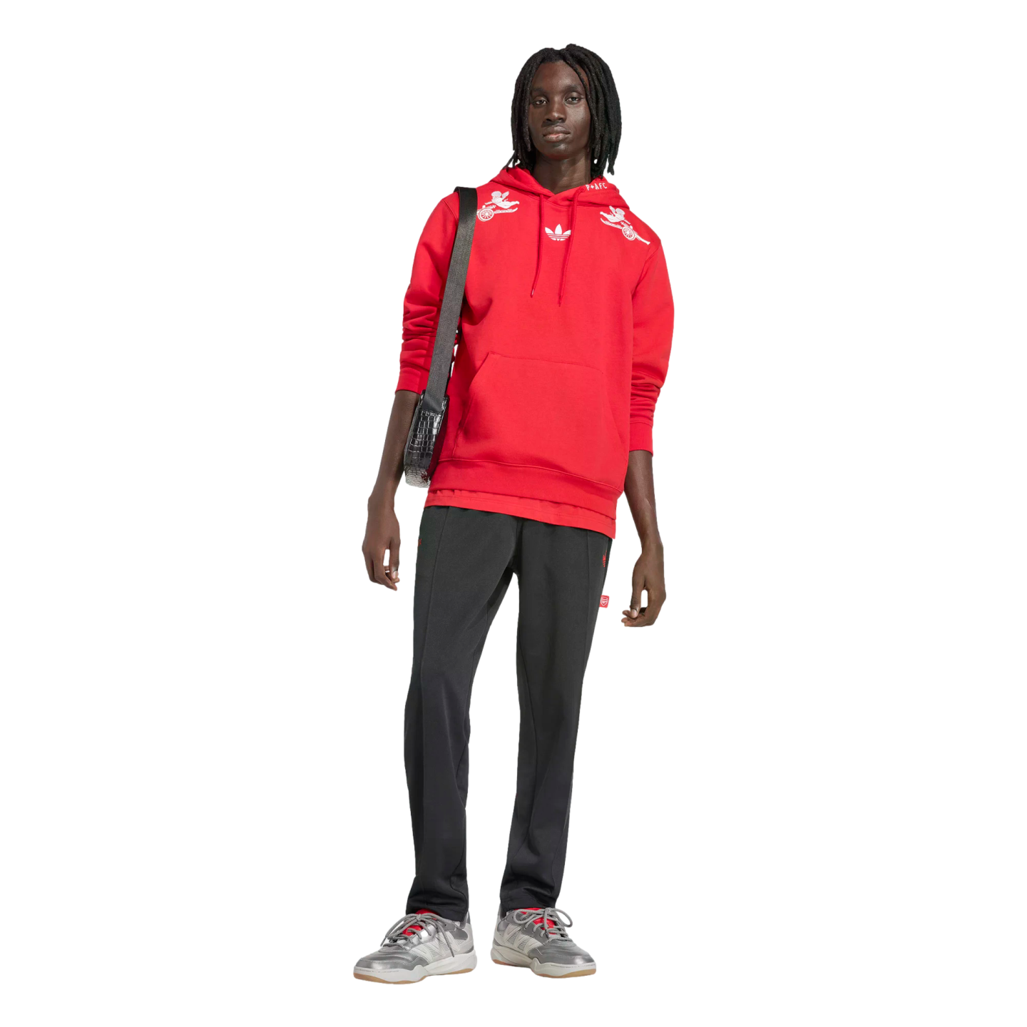 A Black person with dreadlocks stands against a black background in a red hoodie, black track pants, and grey sneakers, with a black crossbody bag.