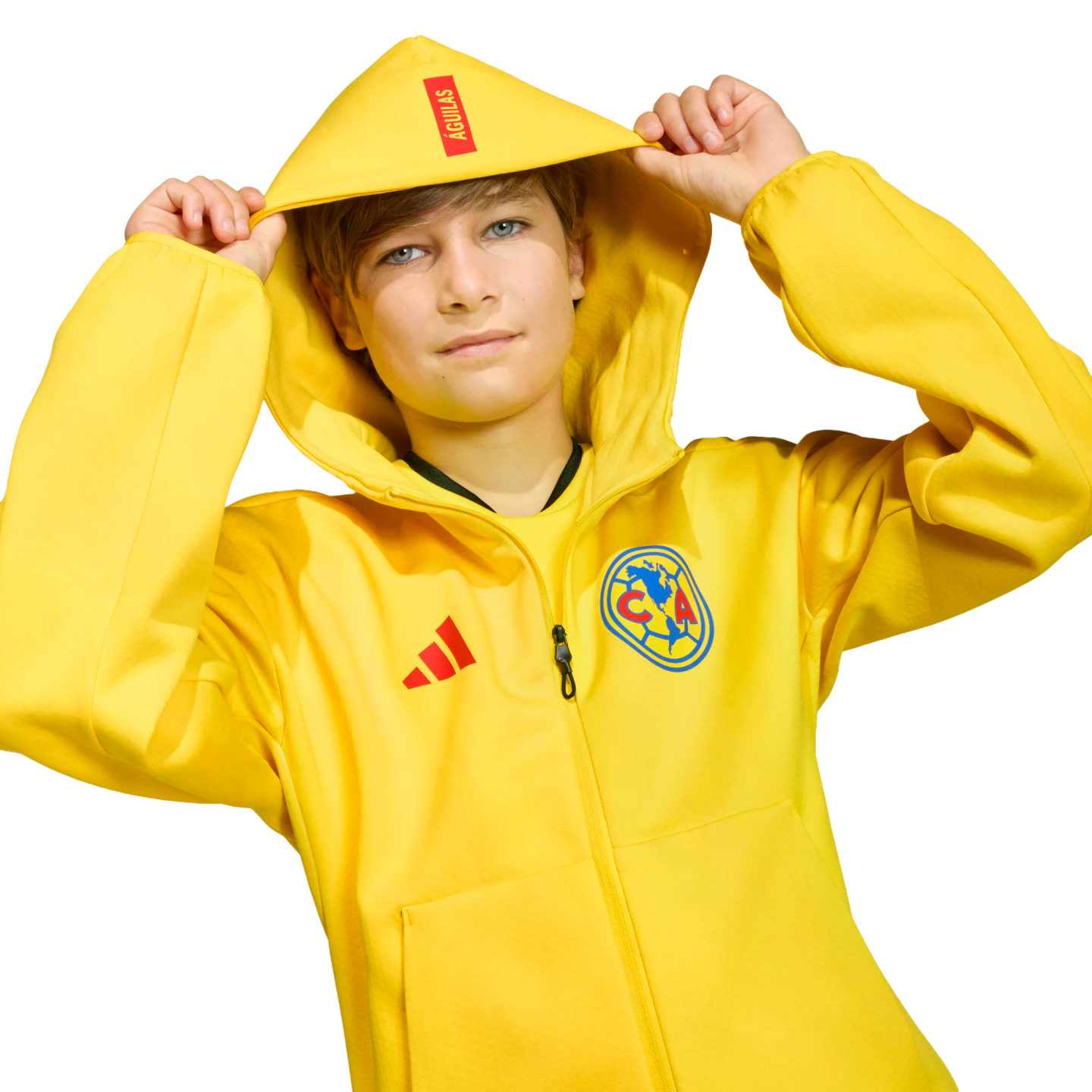 A boy wearing a yellow Adidas Club América hoodie with "ÁGUILAS" on the hood, holding its edges against a black background.