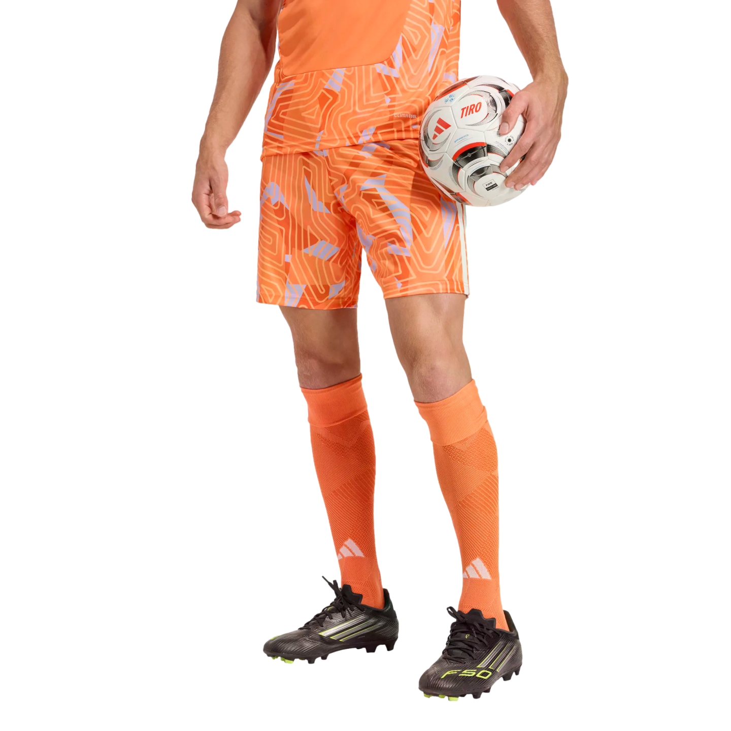 A person from the waist down wearing an orange patterned soccer kit and black cleats, holding a soccer ball against a solid black background.