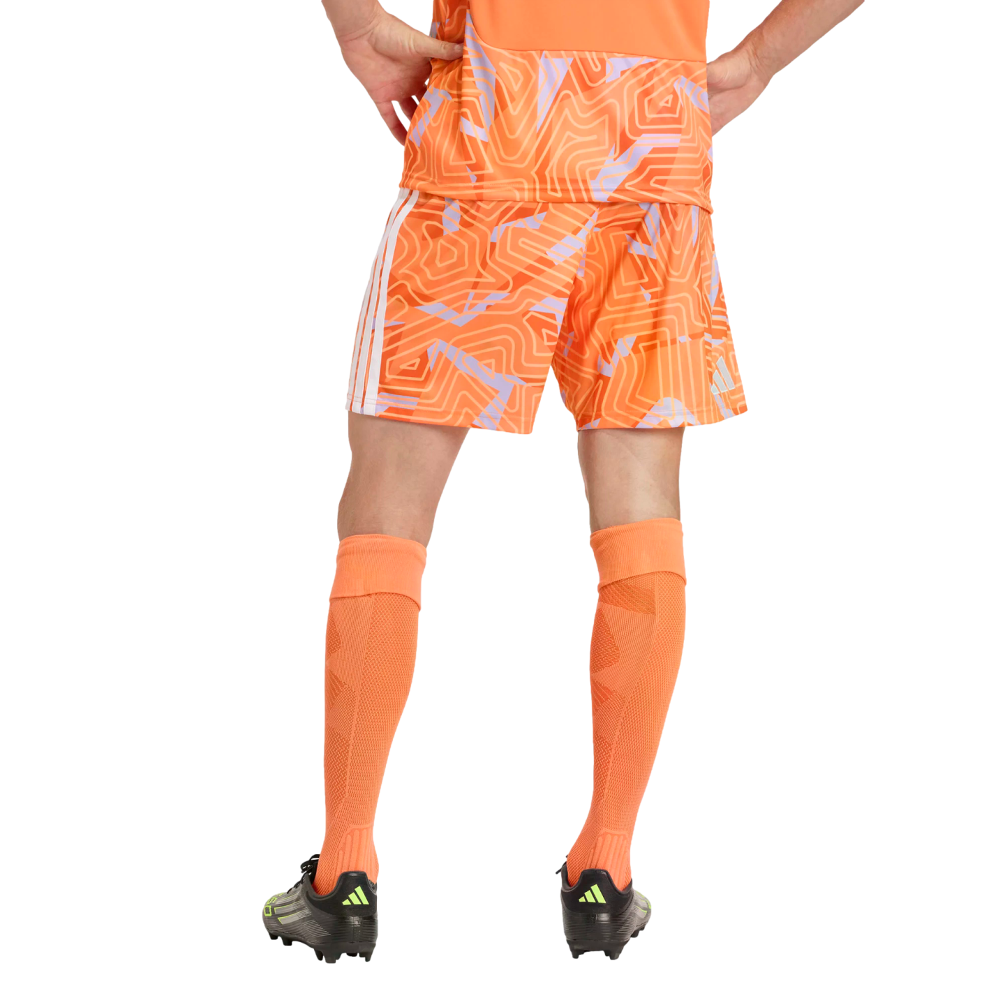 Back view of a person in a patterned orange soccer uniform, tall orange socks, and black cleats, standing with hands on hips against a plain black background.