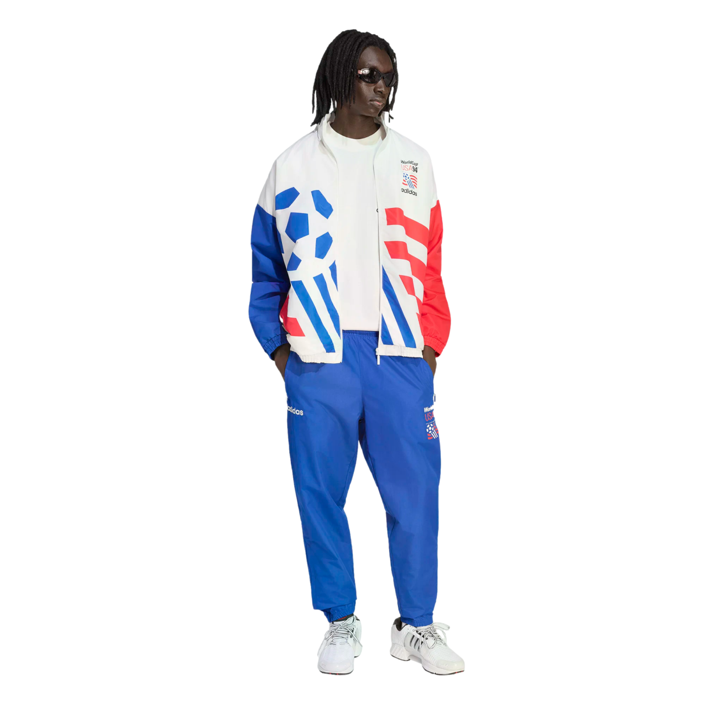 A person models a vintage-style Adidas World Cup USA '94 tracksuit against a black background. The white jacket features bold blue and red graphics, including a soccer ball and stripes, paired with matching blue track pants and white sneakers.