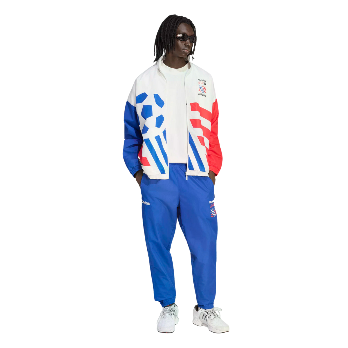 A person models a vintage-style Adidas World Cup USA '94 tracksuit against a black background. The white jacket features bold blue and red graphics, including a soccer ball and stripes, paired with matching blue track pants and white sneakers.