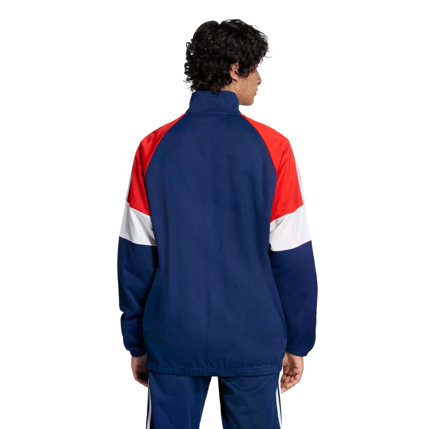 Back view of a man with curly hair wearing a navy, red, and white color-blocked track jacket and matching pants against a black background.