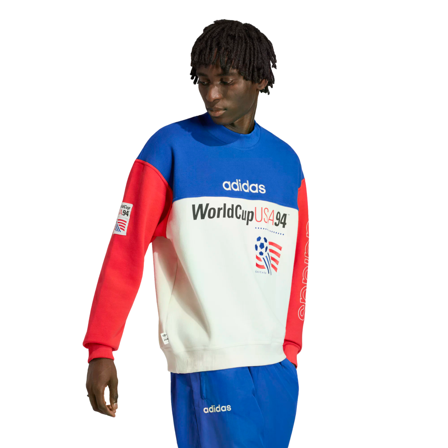 A person with short dreadlocks wearing an Adidas color-blocked "WorldCup USA 94" sweatshirt in blue, white, and red, paired with matching blue track pants, against a solid black background.