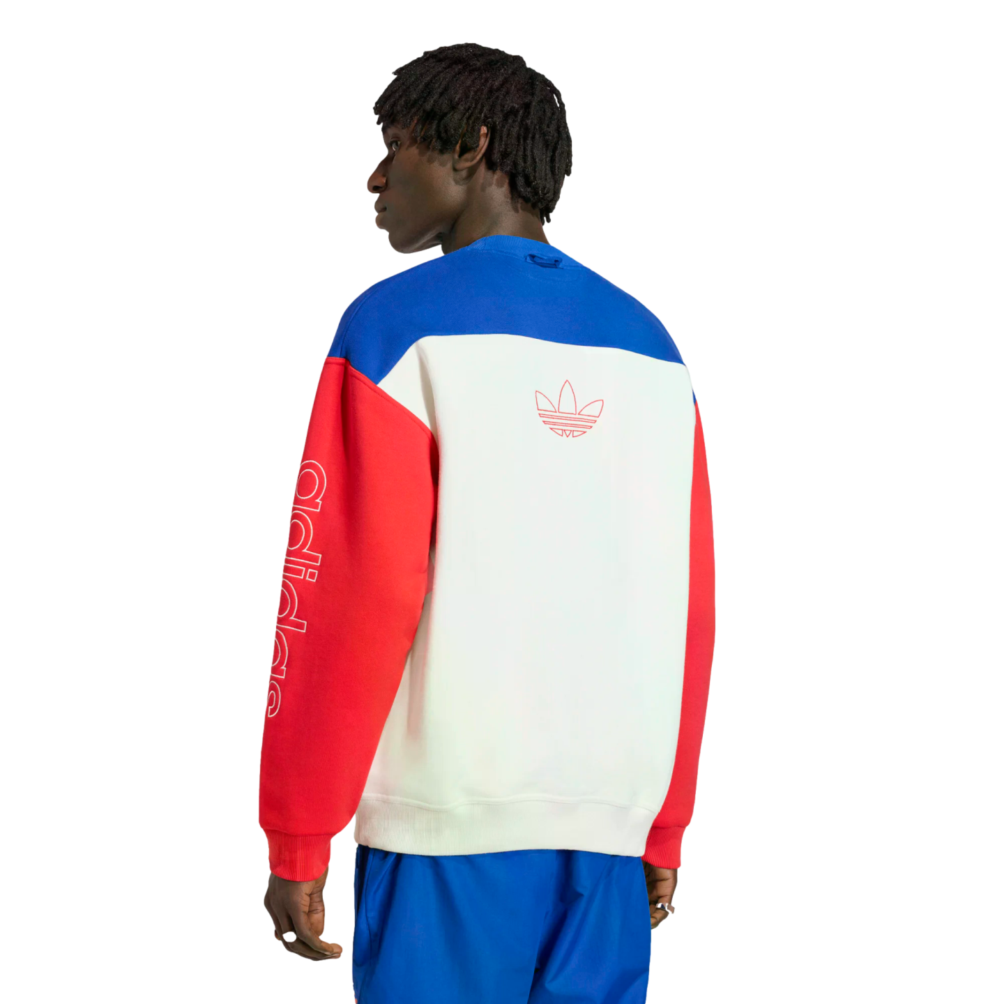 A rear view of a man wearing a color-blocked Adidas crewneck sweatshirt with blue, white, and red panels. The white back panel features a red-outlined Trefoil logo, and the left red sleeve has vertical "adidas" lettering in white. He is standing against a